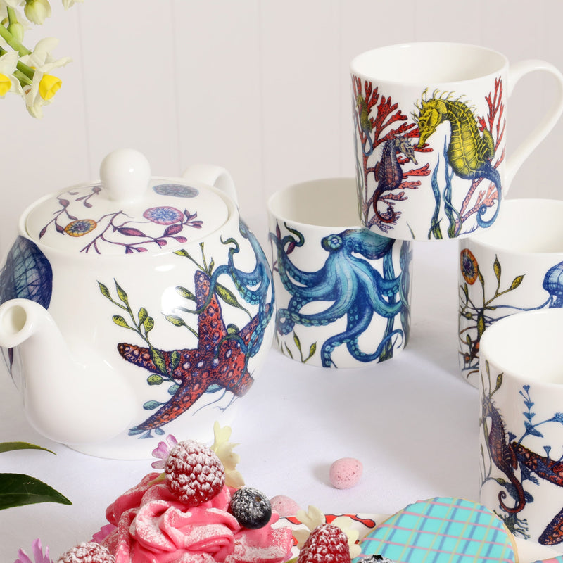 Reef teapot and 4 mugs decorated in our brightly coloured sea creatures and seaweed designs. There are decorated pink cupcakes in the foreground and small soft pink narcissi to the side.