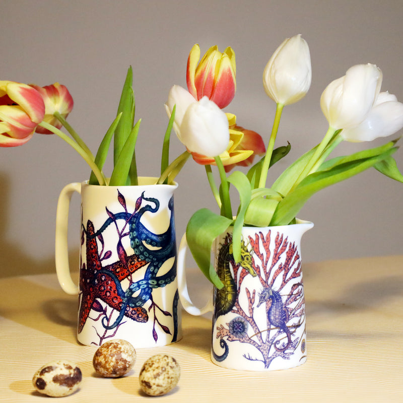2 jugs in our reef design on a striped yellow table cloth and 3. quails eggs in the foreground. The jugs are decorated on brightly coloured sea creatures octopus, starfish seahorse, corals and seaweeds. The jugs are filled with red and yellow tulips and white tulips.