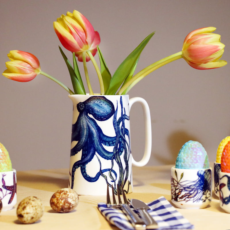 White jug with brightly coloured blue and green octopus with red and yellow tulips in. this is sitting on a yellow table with the same designed egg cups with sequined eggs in. 