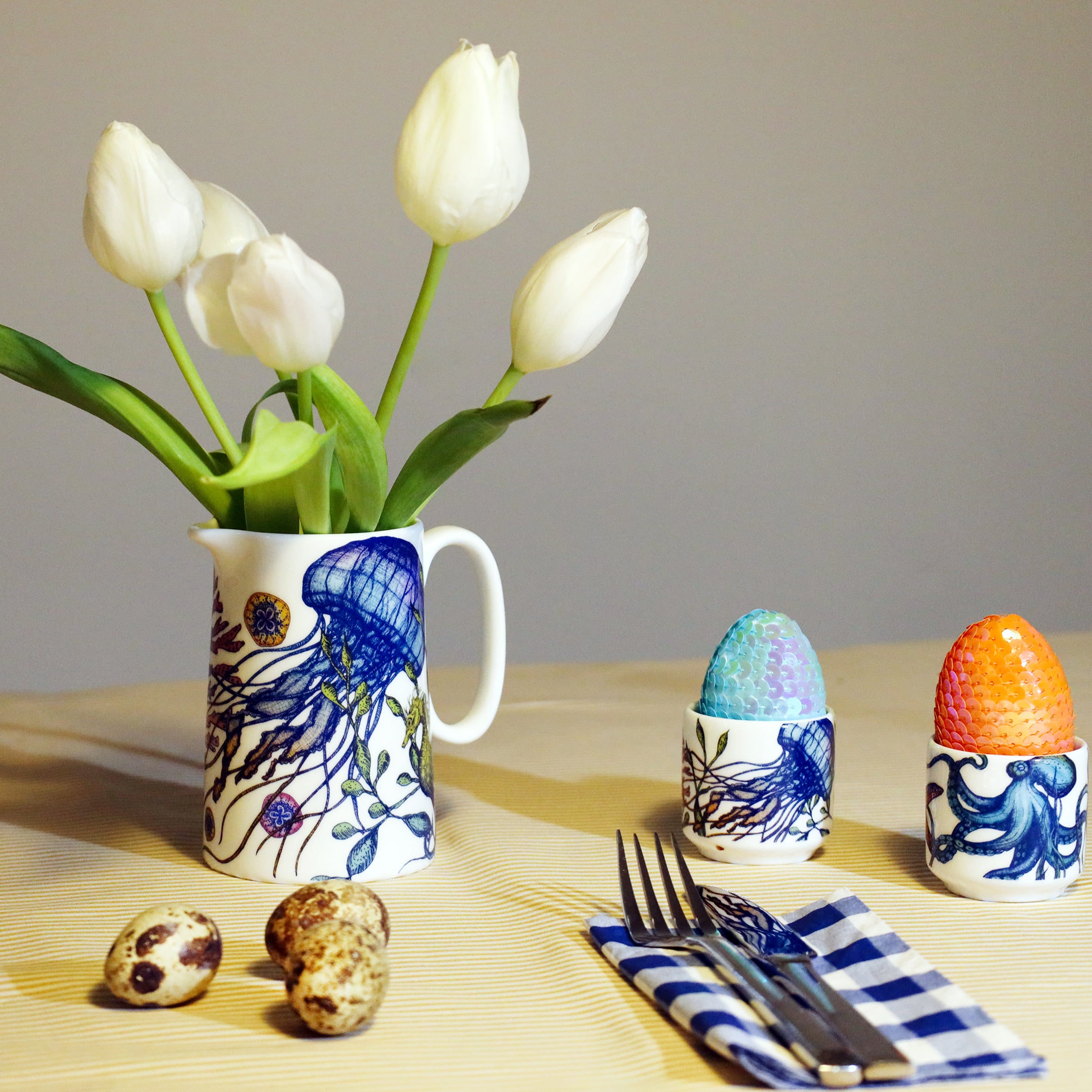 White jug with brightly coloured jellyfish and seaweed design filled with white tulips and sitting on a yellow striped tablecloth. There are 2 egg cups from the same reef range to the sidde with an octopus on one and a jellyfish on the other and they each have a sequined egg in them. There are 3 quails eggs to the front and a folded blue gingham napkin with a knife and fork.