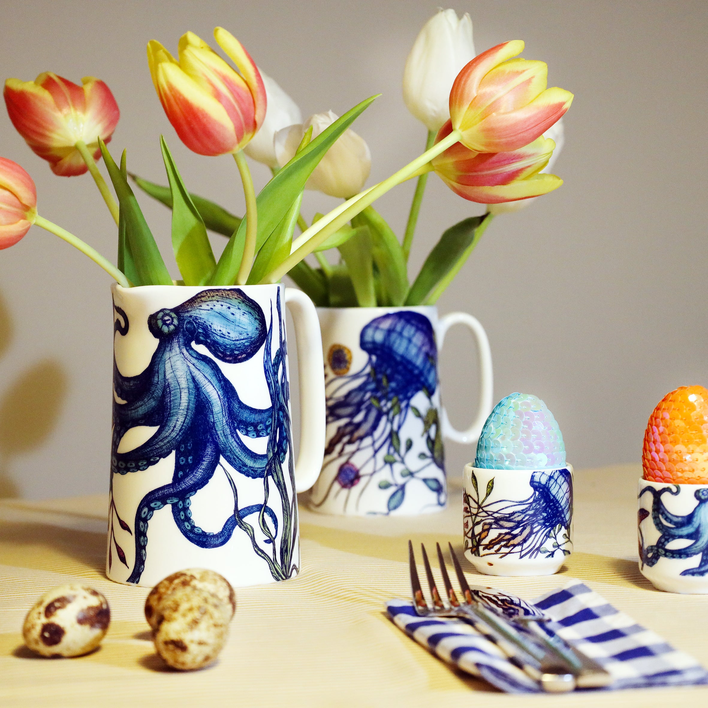 White jug with brightly coloured blue and green octopus with red and yellow tulips in. this is sitting on a yellow table with the same designed egg cups with sequined eggs in. There is a smaller jug from the same collection with a jellyfish on it in the background as well.