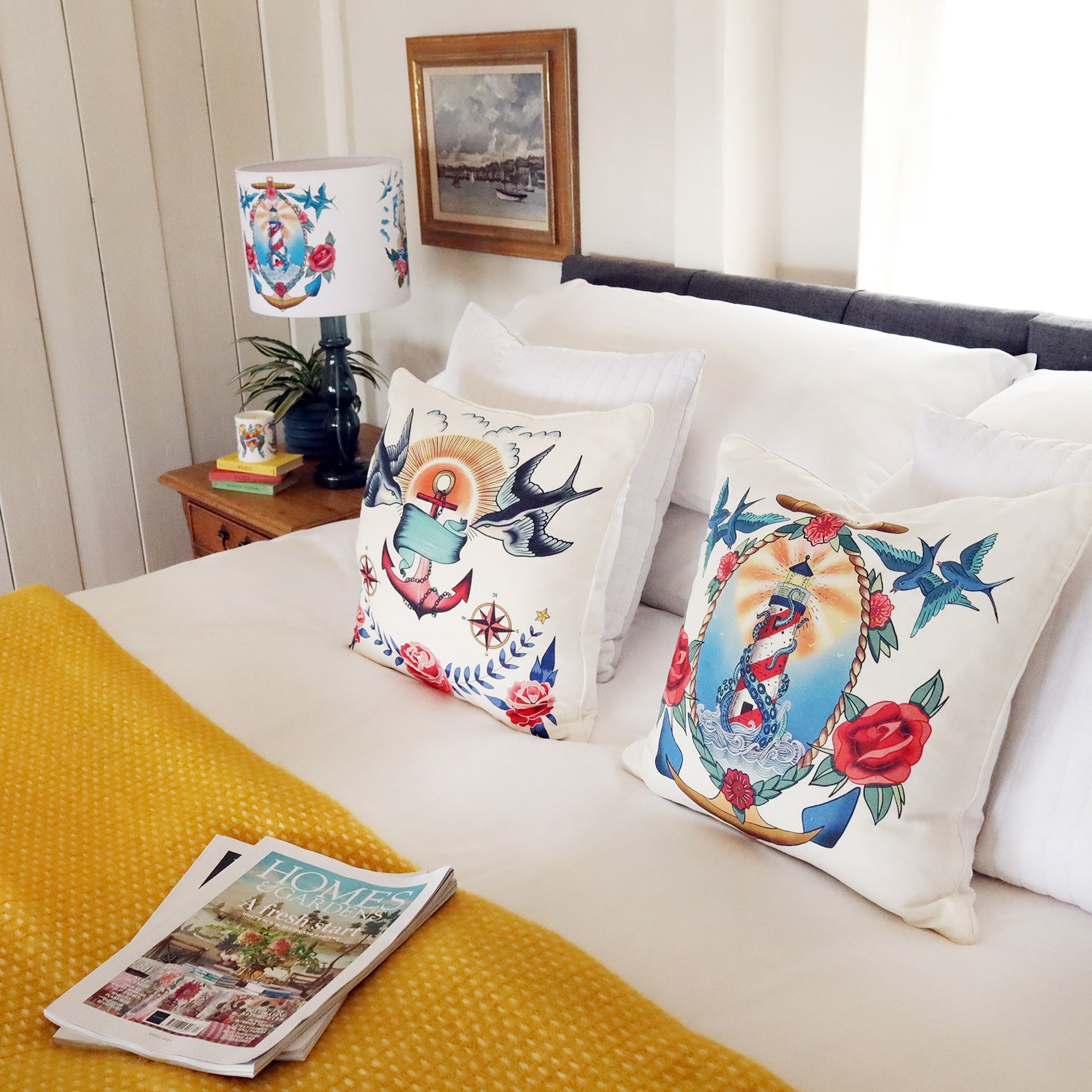 A bedroom scene with 2 white cushions propped up against the pillows each with a brightly coloured tattoo inspired design on . There is a yellow throw on the bed and a matching tattoo lampshade and bone china candle to the side.