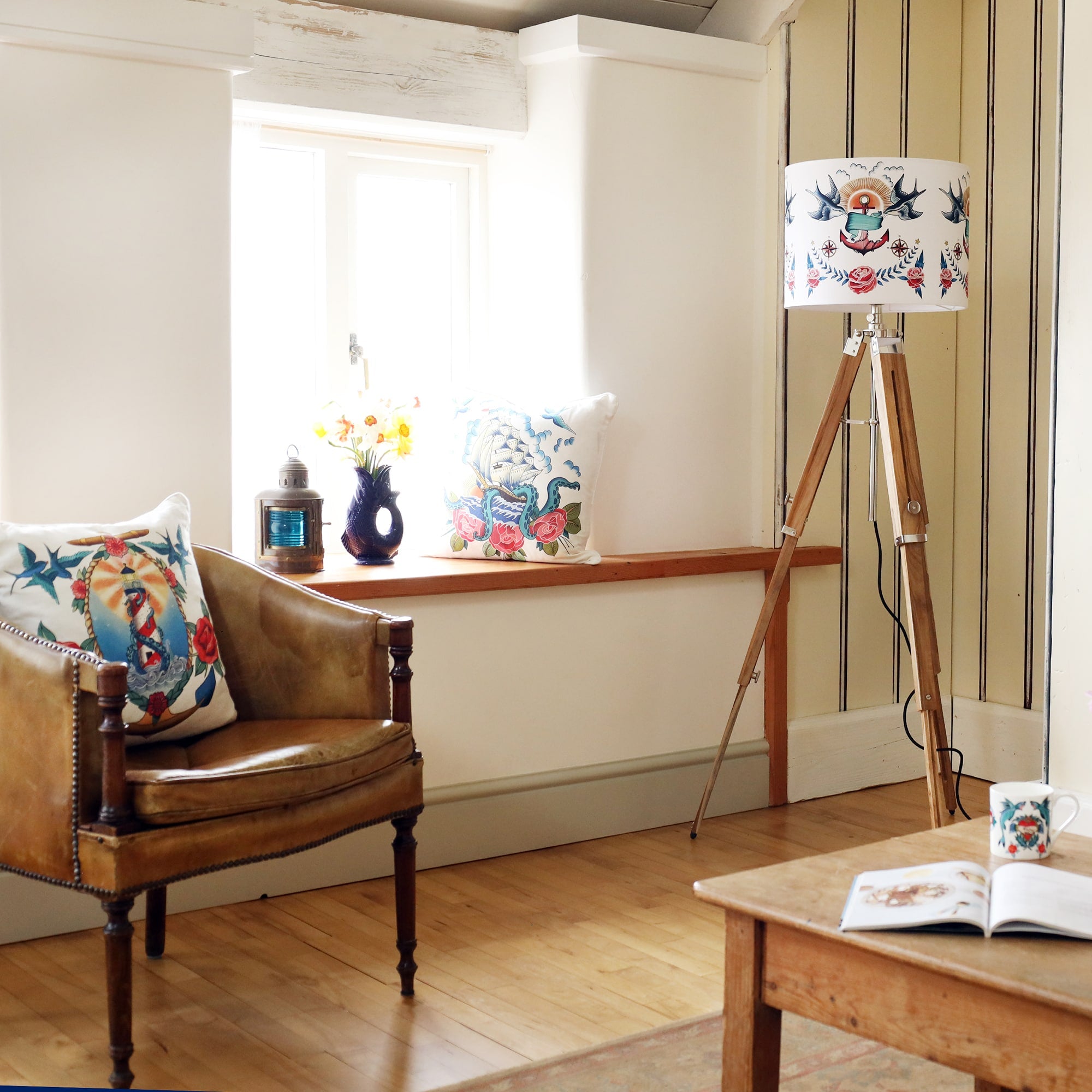 Room setting with wooden tripod lamb base with sailor's tattoo inspired lampshade on it. There is a cushion on the window seat with a vase of flowers and a ships lantern and on the chair in the foreground is a cushion with a tattoo inspired design of lighthouse and kraken.