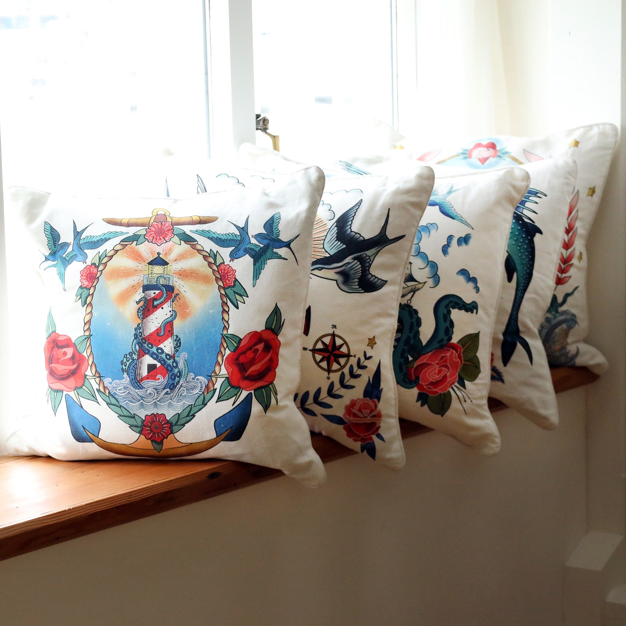 Row of white cushions on a sunny window ledge all with a different brightly coloured sailor's tattoo inspired design on them.