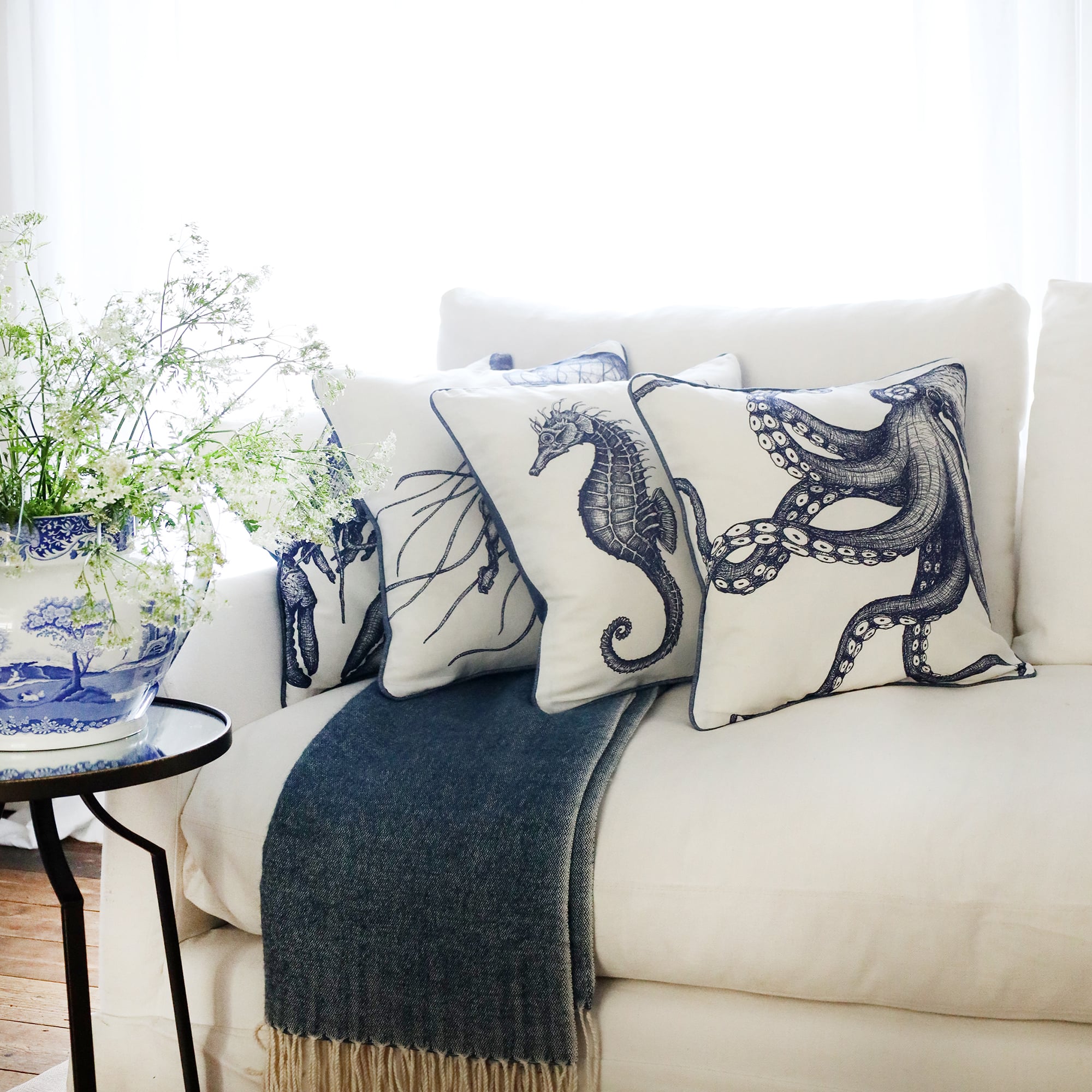 row of 4 blue & white illustrated with various sea creatures cushions and a blue & white octopus cushion at the front, sitting on a navy throw on a white sofa with bright sunlight window behind and a large willow pattern jug filled with cow parsley