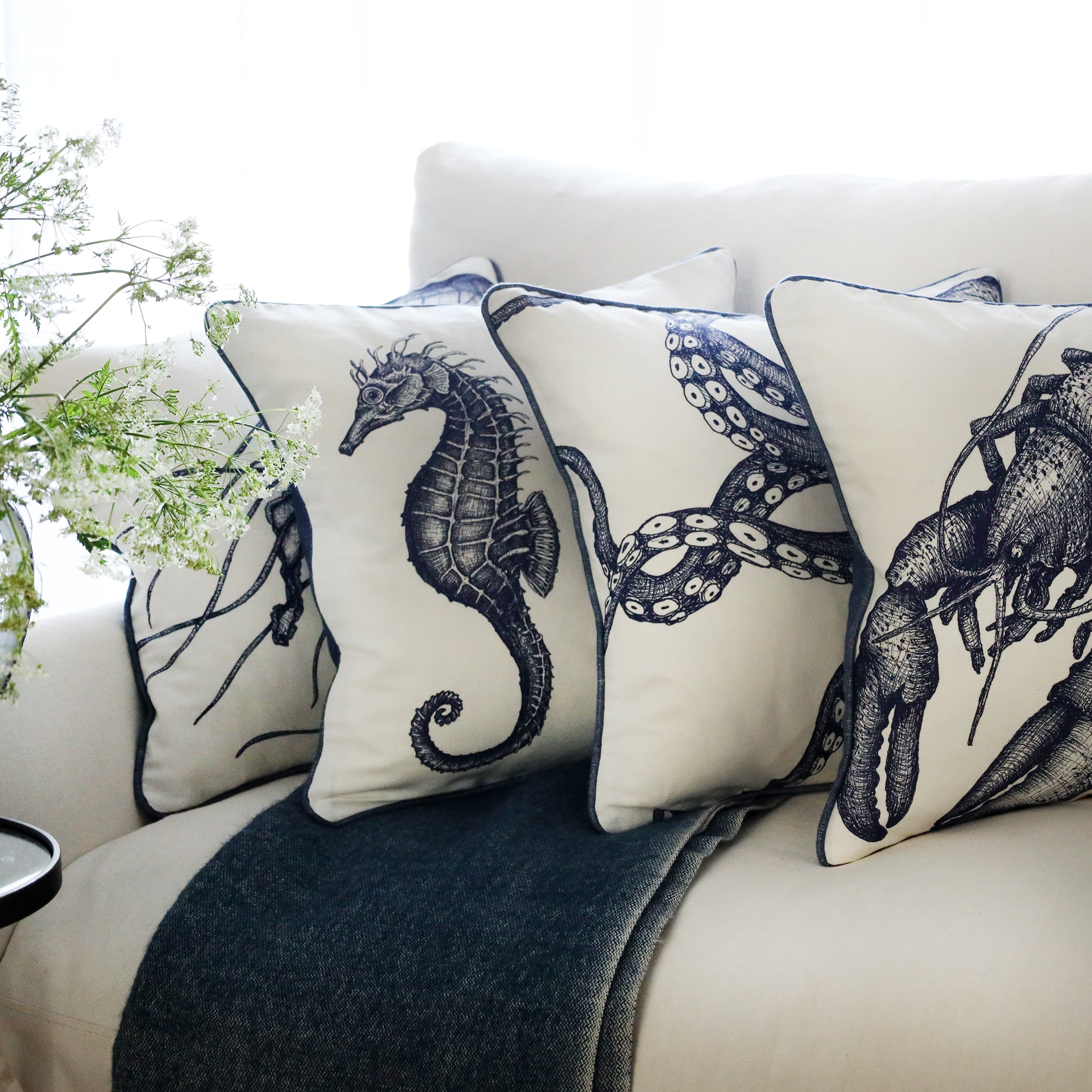 row of 4 blue & white illustrated sea creature cushions with a lobster cushion at the front, sitting on a navy throw on a white sofa with bright sunlight window behind and a large willow pattern jug filled with cow parsley