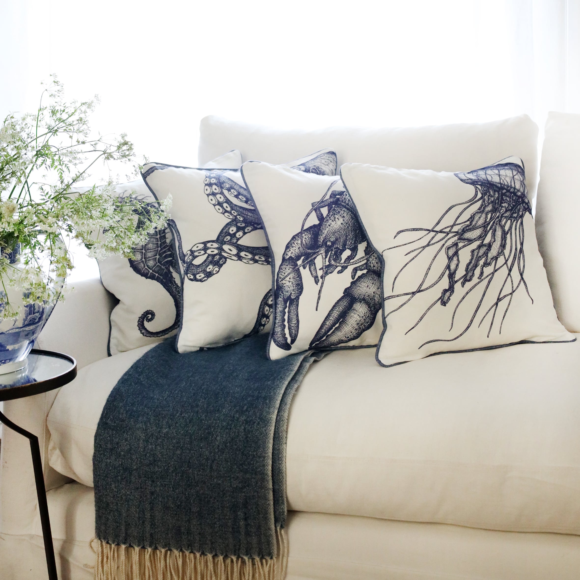 row of 4 blue & white illustrated sea creature cushions with a jellyfish cushion at the front, sitting on a navy throw on a white sofa with bright sunlight window behind and a large willow pattern jug filled with cow parsley