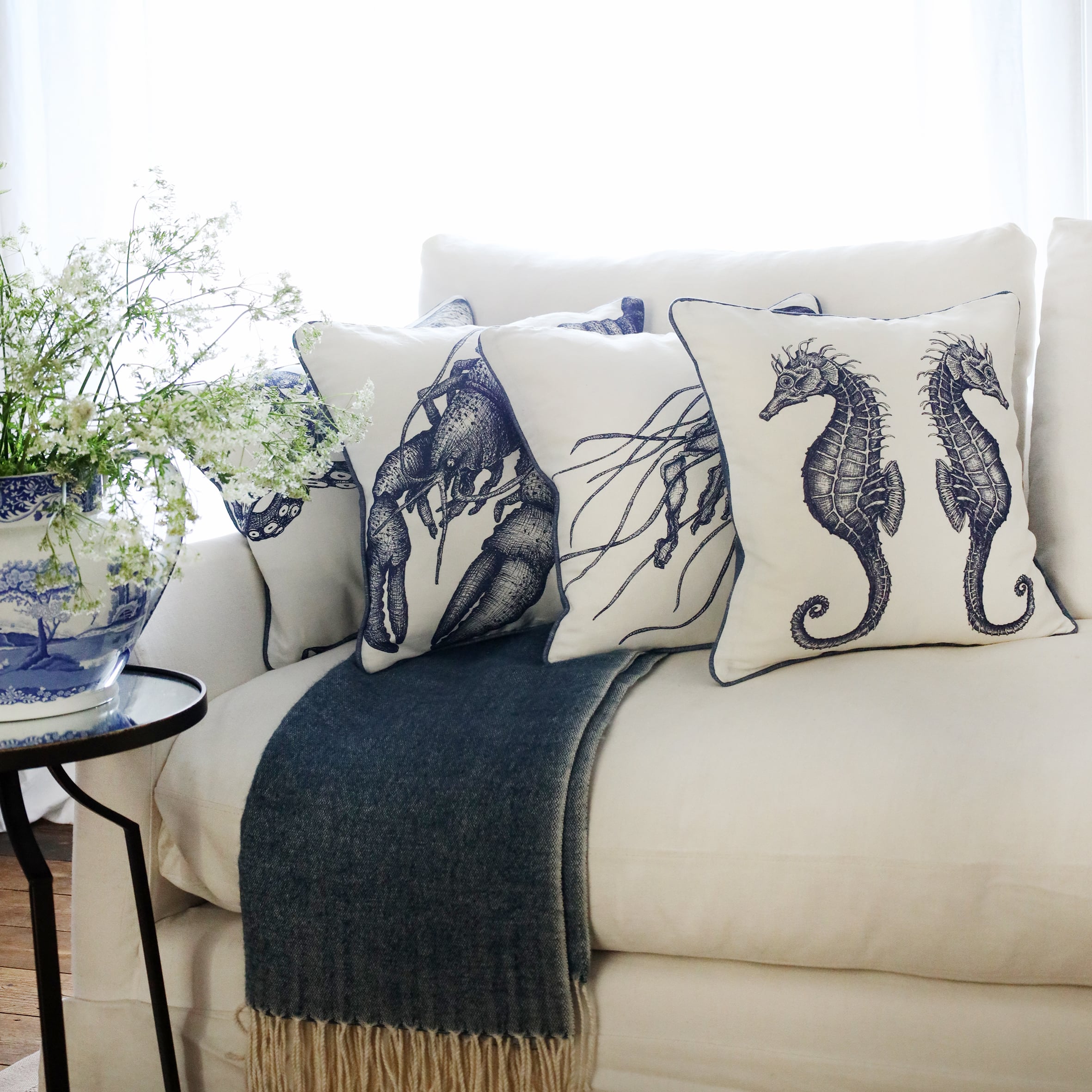row of 4 blue & white illustrated sea creature cushions with a seahorse cushion at the front, sitting on a navy throw on a white sofa with bright sunlight window behind and a large willow pattern jug filled with cow parsley