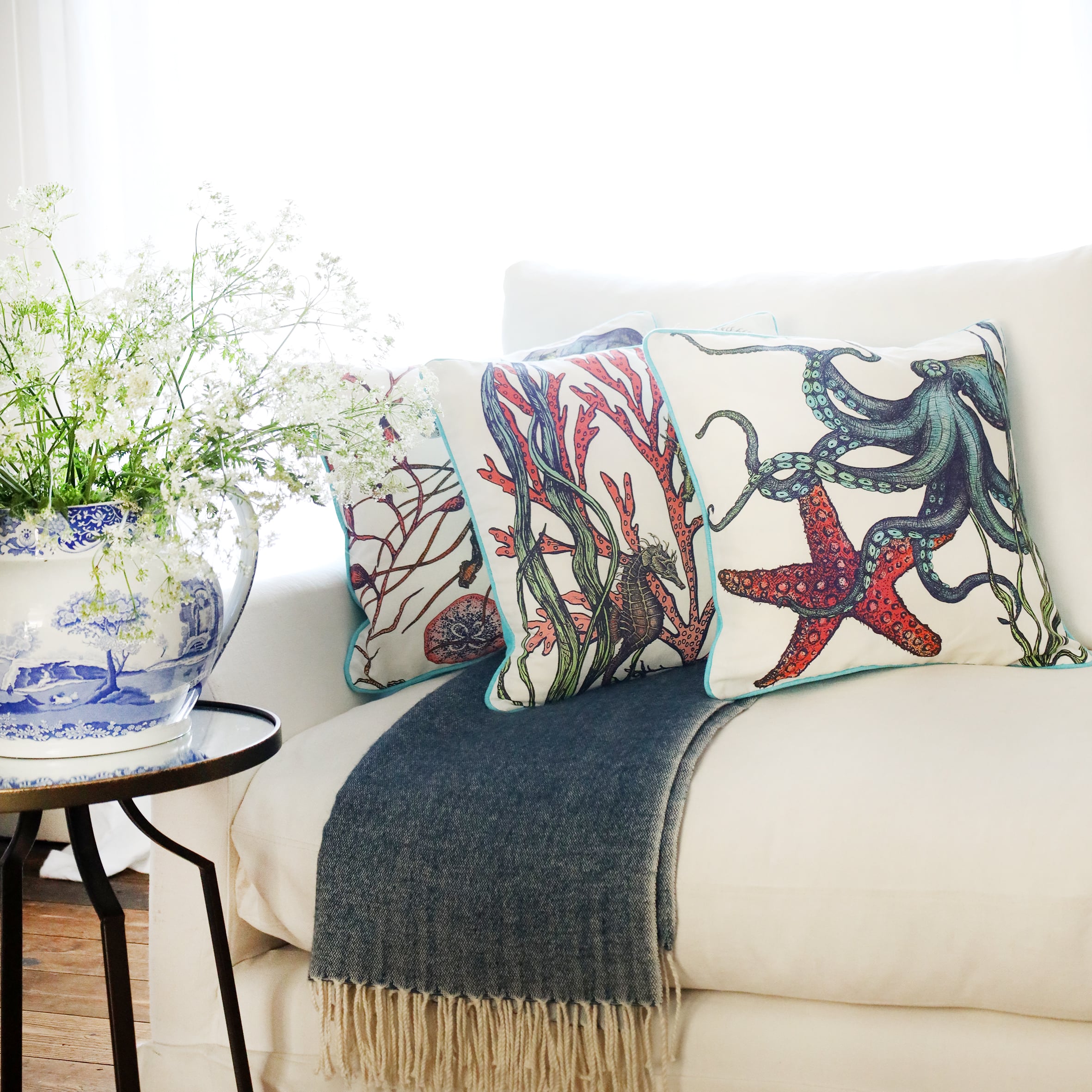 3 cushions an a white sofa with brightly coloured sea creatures illustrated  on with the sun streaming through the window at the back and a large willow pattern jug filled with cow parsley