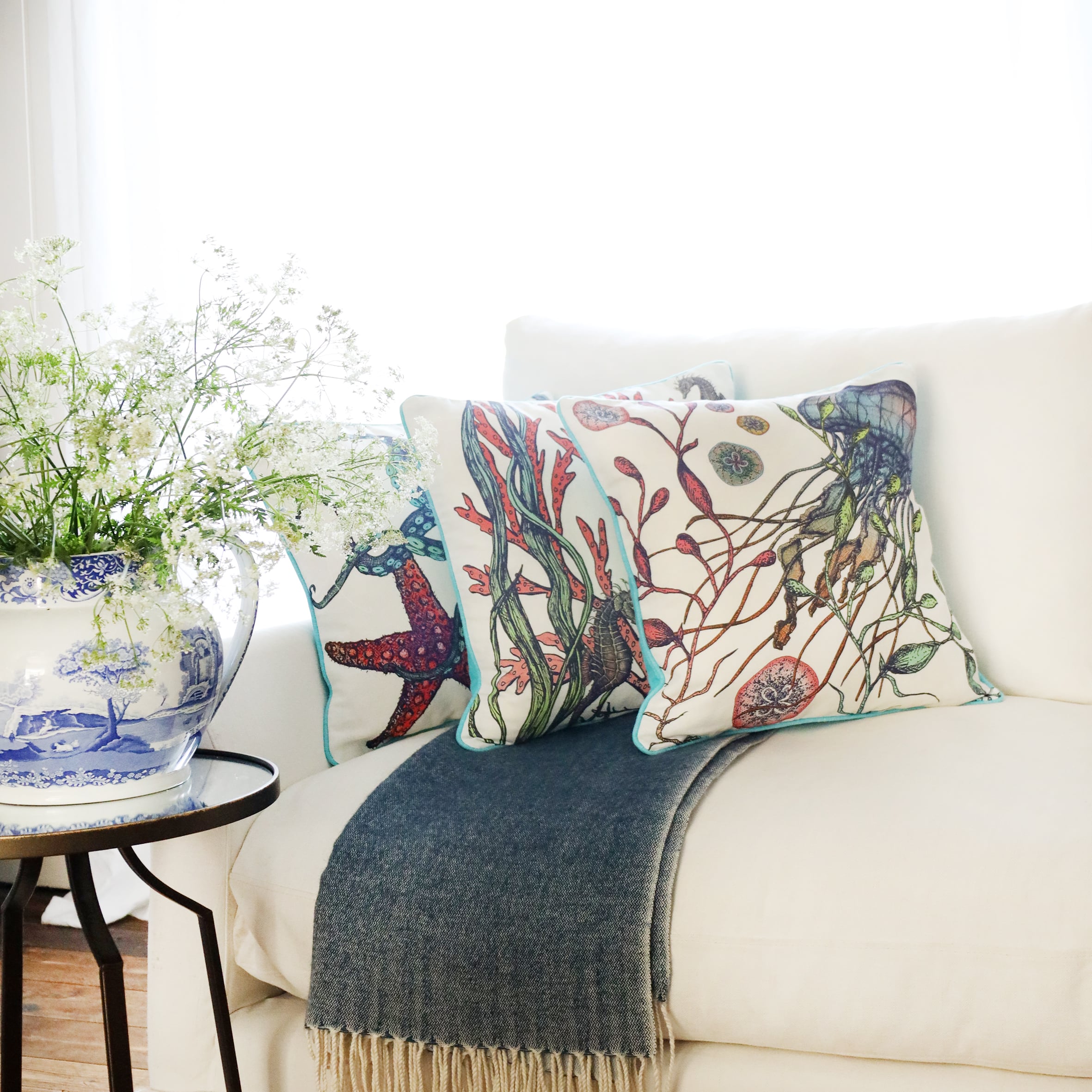 3 cushions an a white sofa with brightly coloured sea creatures illustrated  on with the sun streaming through the window at the back and a large willow pattern jug filled with cow parsley