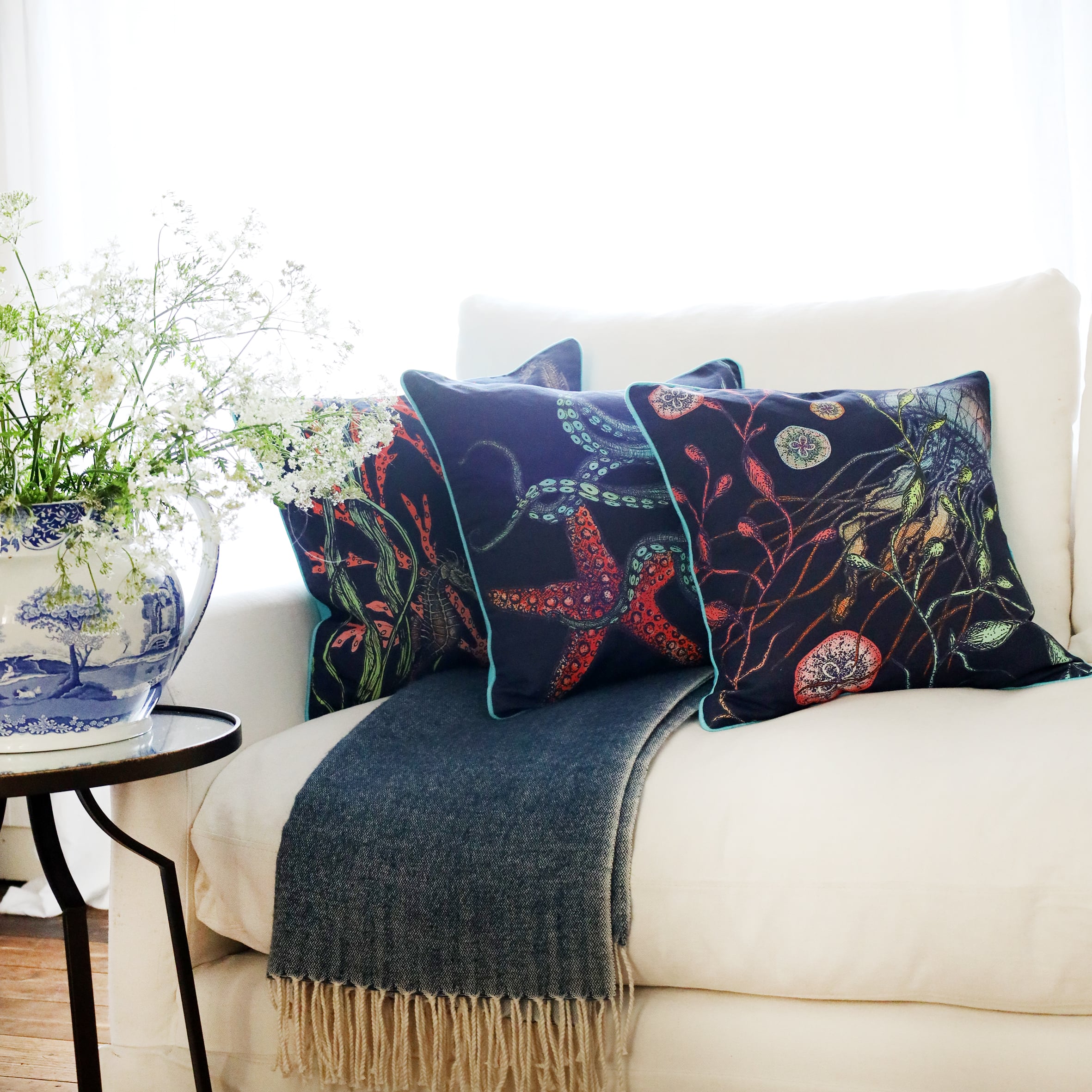 3 cushions an a white sofa with brightly coloured sea creatures illustrations on a navy ground with the sun streaming through the window at the back and a large willow pattern jug filled with cow parsley