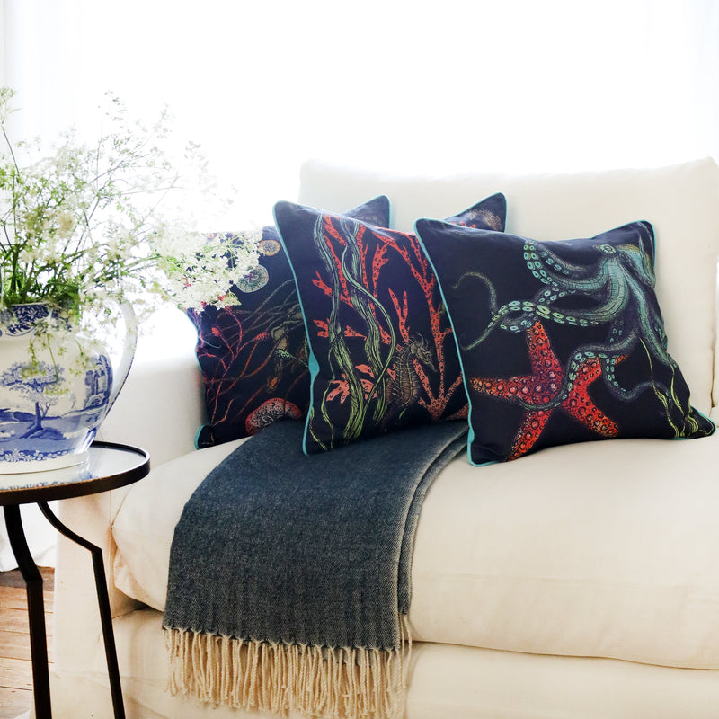 3 cushions an a white sofa with brightly coloured sea creature illustrations on a navy ground, with the sun streaming through the window at the back and a large willow pattern jug filled with cow parsley