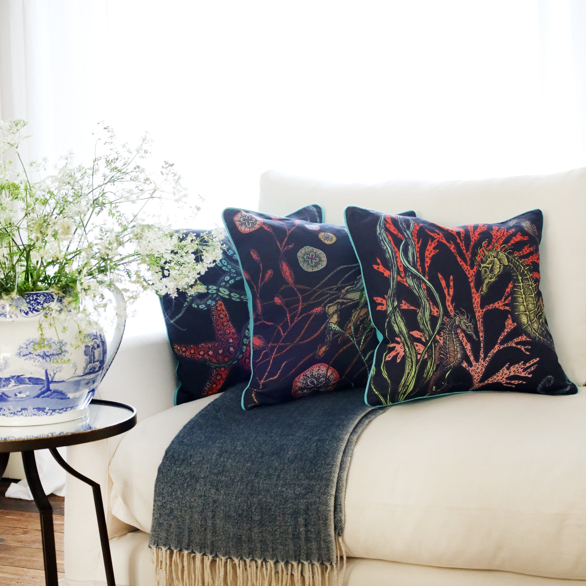 3 cushions an a white sofa with brightly coloured sea creatures illustrated on a navy ground with the sun streaming through the window at the back and a large willow pattern jug filled with cow parsley