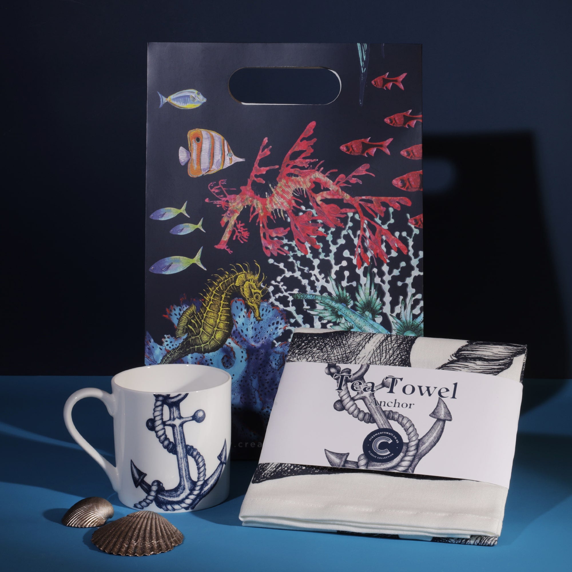 A nautical-themed gift set featuring a mug, tea towel, and a gift bag. The mug and tea towel are decorated with blue anchor illustrations, while the gift bag showcases a colorful underwater scene with fish, coral, and a seahorse. Decorative seashells are placed alongside the items, enhancing the coastal theme against a dark blue background.