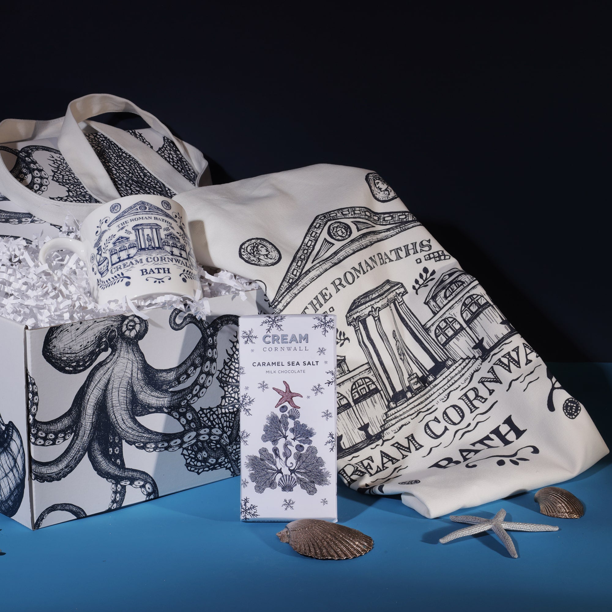 A Roman Baths-themed gift set from Cream Cornwall, featuring a mug, tote bag, and a bar of caramel sea salt chocolate. The mug and tote bag are decorated with intricate illustrations of the Roman Baths in blue. The items are presented in a box adorned with an octopus design, accompanied by decorative seashells and starfish, enhancing the coastal aesthetic against a dark blue background.