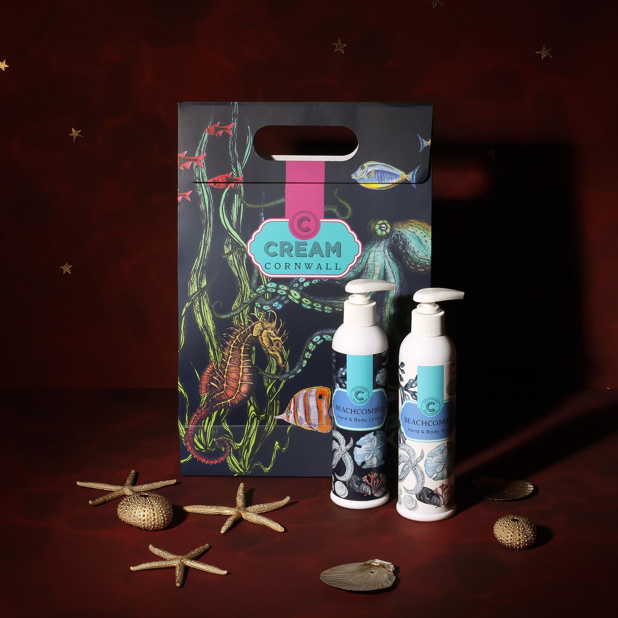 A Cream Cornwall Beachcomber-themed bath gift set, featuring hand and body lotion, and body wash bottles. The items are displayed with a colorful gift bag decorated with marine life illustrations, including fish, seaweed, and a seahorse. Decorative starfish and seashells are scattered around the products, enhancing the coastal theme against a deep red background.