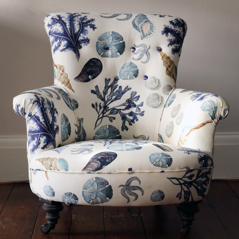 Front view of a  re upholsterd victorian button back armchair covered in a shell and seaweed fabric in blue tones on an off white background.