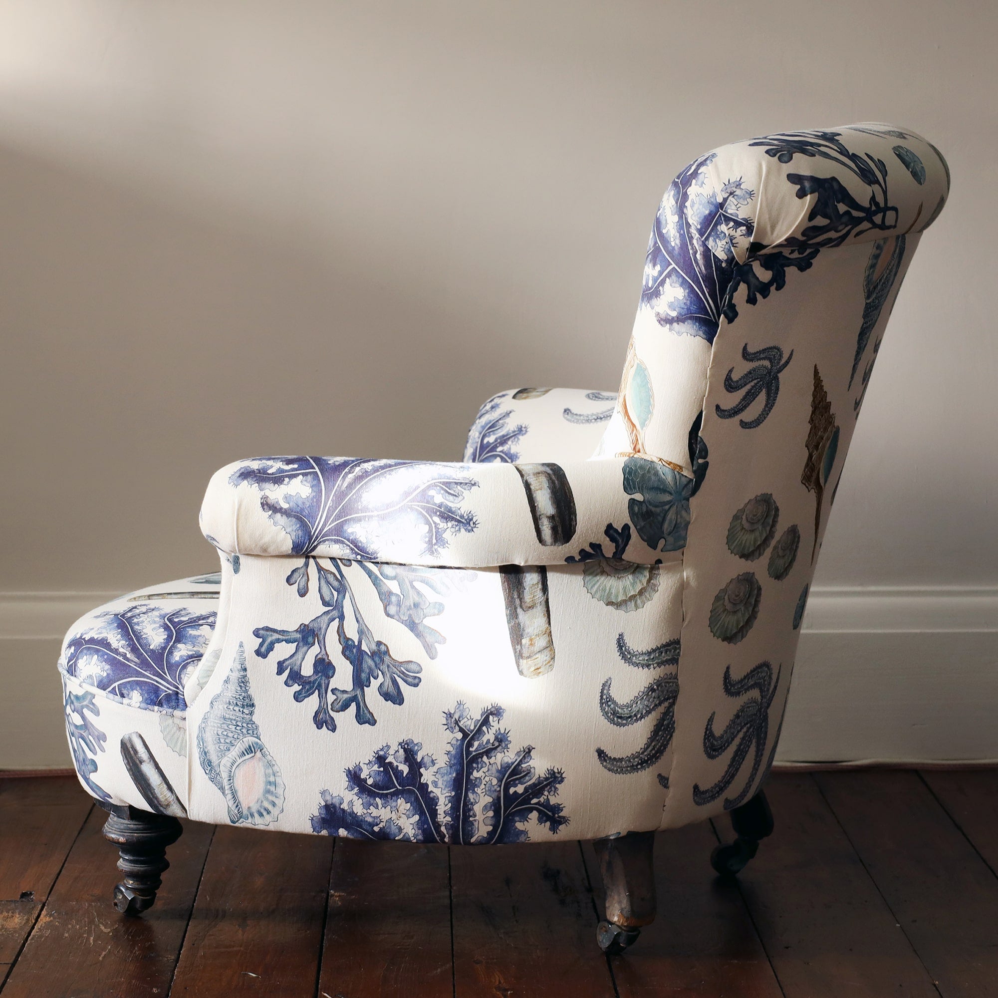 Side view of re upholstered victorian button back armchair covered in a shell and seaweed fabric in blue tones on an off white background.