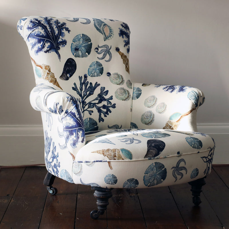 Side angled view of re upholstered victorian button back armchair covered in a shell and seaweed fabric in blue tones on an off white background.