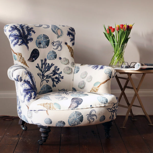 A reupholsterd victorian button back armchair covered in a shell and seaweed fabric, sitting next to a small table with a vase of red tulips a folded magazine and reading glasses.