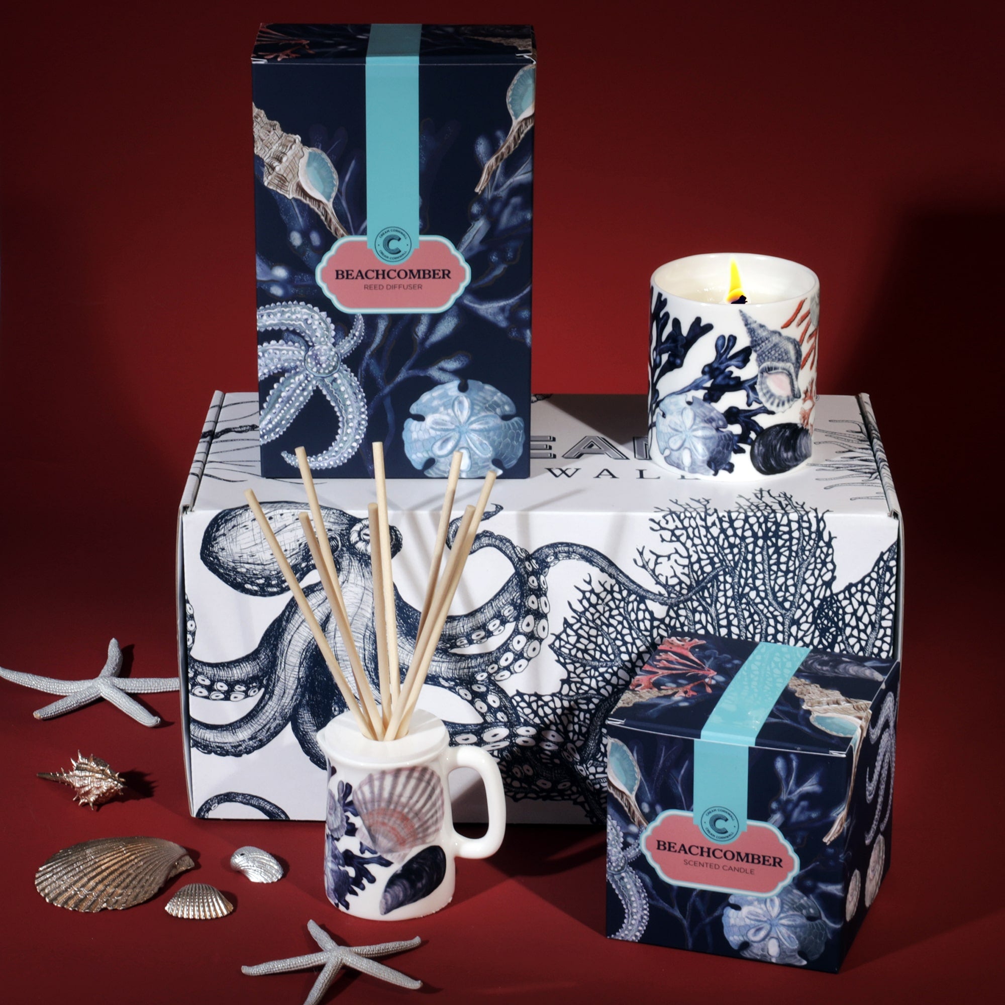 A Cream Cornwall Beachcomber-themed fragrance gift set, featuring a reed diffuser and a scented candle. The packaging and candle holder are decorated with ocean-inspired illustrations of seashells, starfish, and coral in navy and white tones. The products are displayed on a box with an octopus design, accompanied by decorative seashells and starfish, against a deep red background, emphasizing the coastal theme.