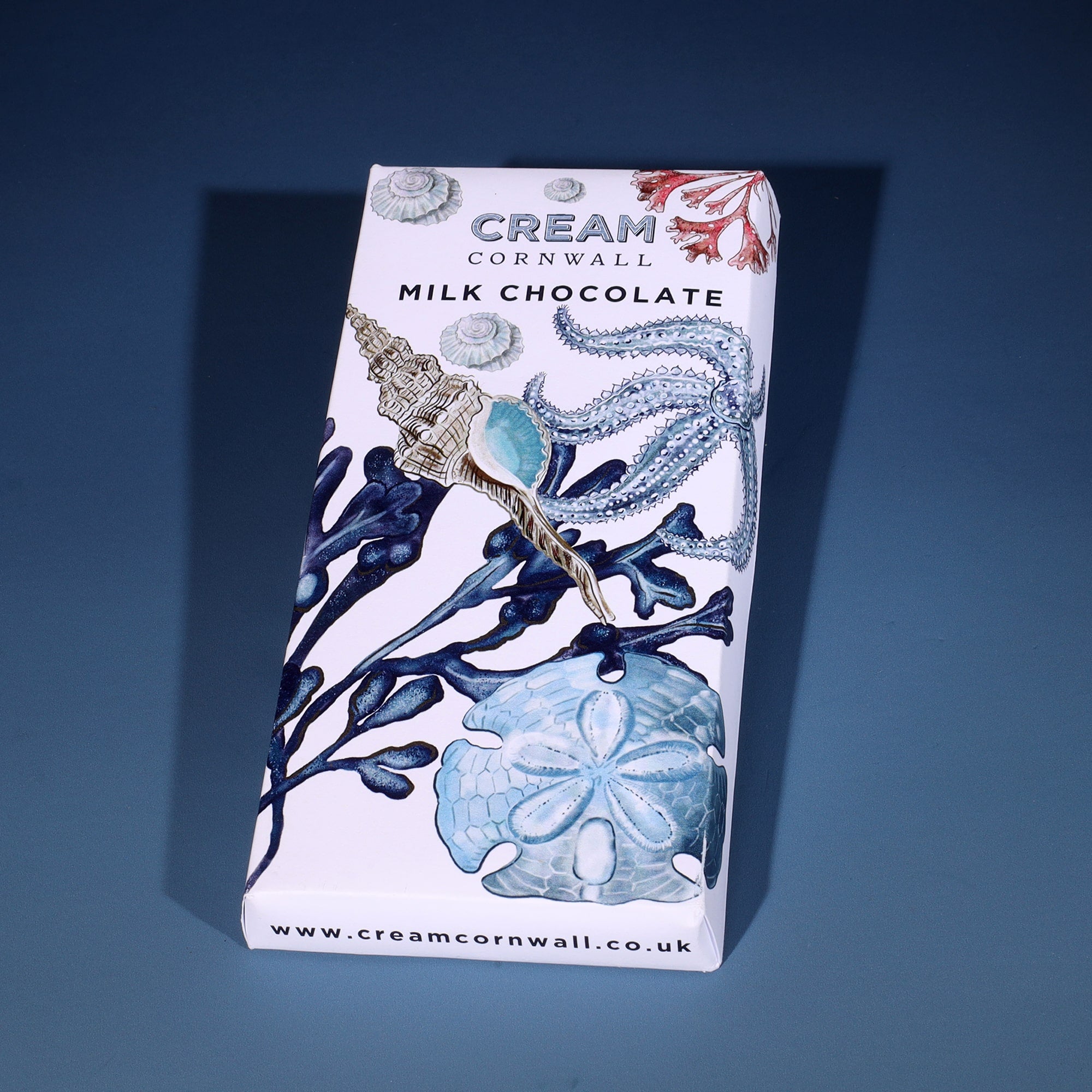 Milk chocolate bar wrapped in a beachcomber themed wrapper with sea sheels, seaweed, starfish & sand dollar. The chocolate bar has been photographed on a graduated blue background.