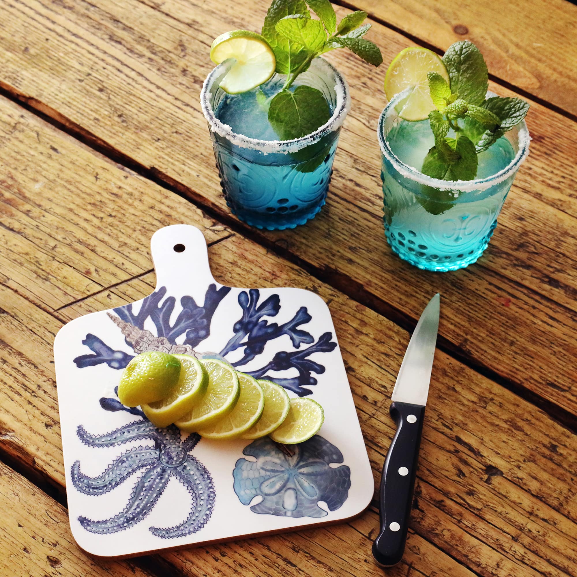 Beachcomber Chopping Board placed on a wooden table.On the board is a sliced lime next to a knife,also on the table are filled blue coloured glasses