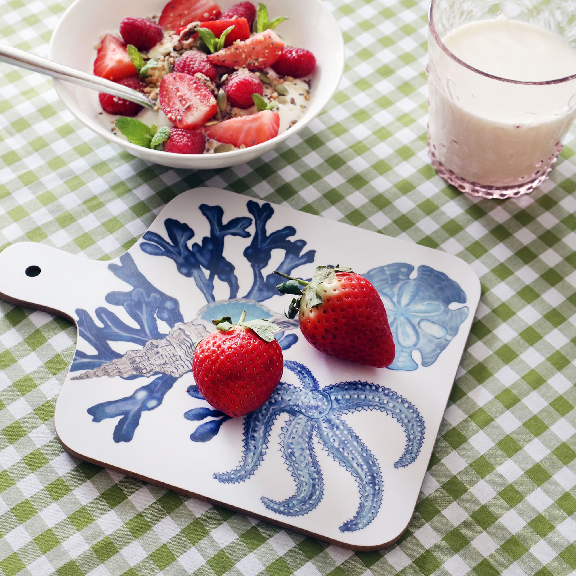 Beachcomber Chopping Board on a green gingham cloth.Also on the table is a bowl with strawberries and seeds next to a glass of milk in a pink tumbler