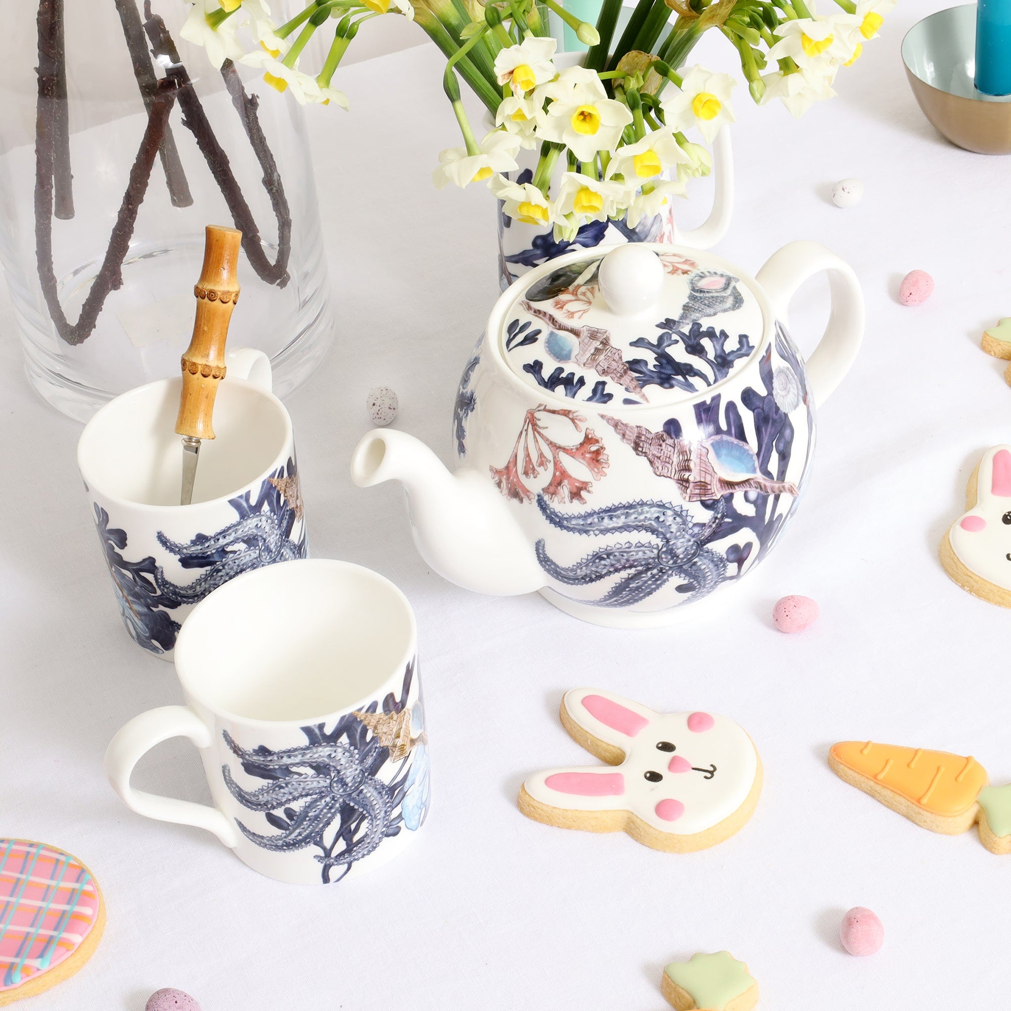 Beachcomber teapot and 2 mugs decorated in hand drawn shells and seaweed in blues and soft pinks. These are on a table with easter themed biscuits, mini candy coloured eggs and spring flowers.