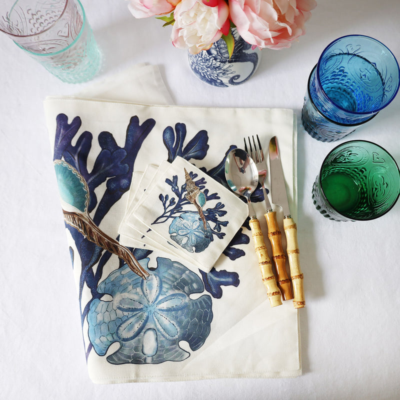 Fabric coasters in our Beachcomber design in a set of four,placed on the matching placemats.Also on the table are bamboo cutlery and some coloured glasses and a seahorse vase with flowers