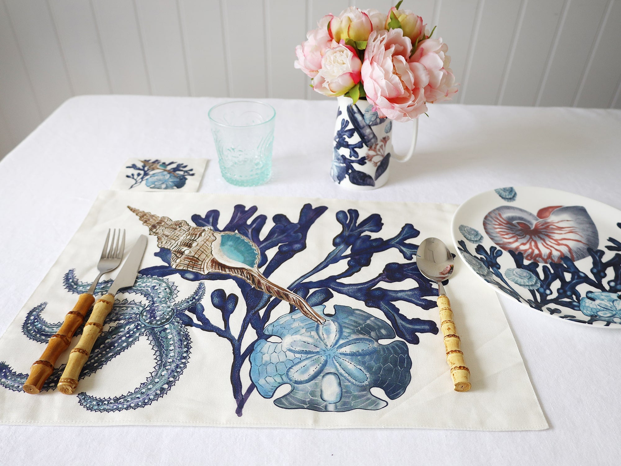 Fabric coaster in our Beachcomber design placed next to a matching placemat on a tablecloth.Also you can see bamboo cutlery,A vase of flowers and a beachcomber bowl and a glass