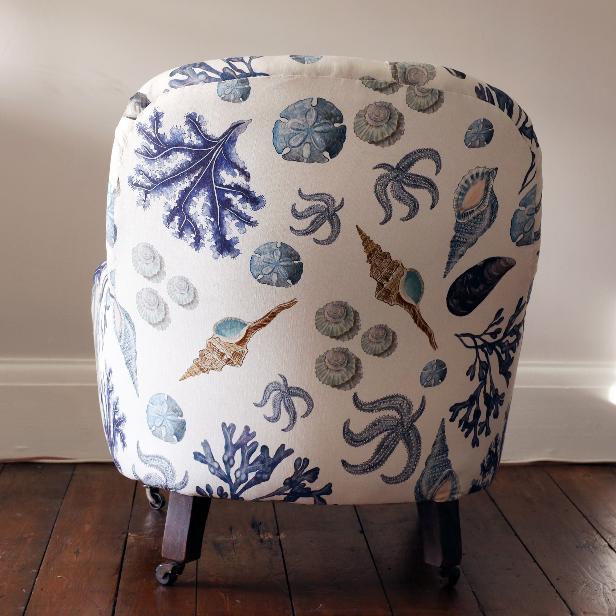 Back view of a re upholsterd victorian button back armchair covered in a shell and seaweed fabric in blue tones on an off white background.