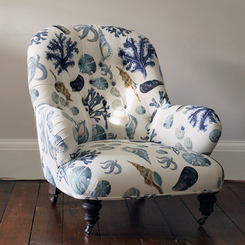 Front view of a re upholsterd victorian button back armchair covered in a shell and seaweed fabric in blue tones on an off white background.