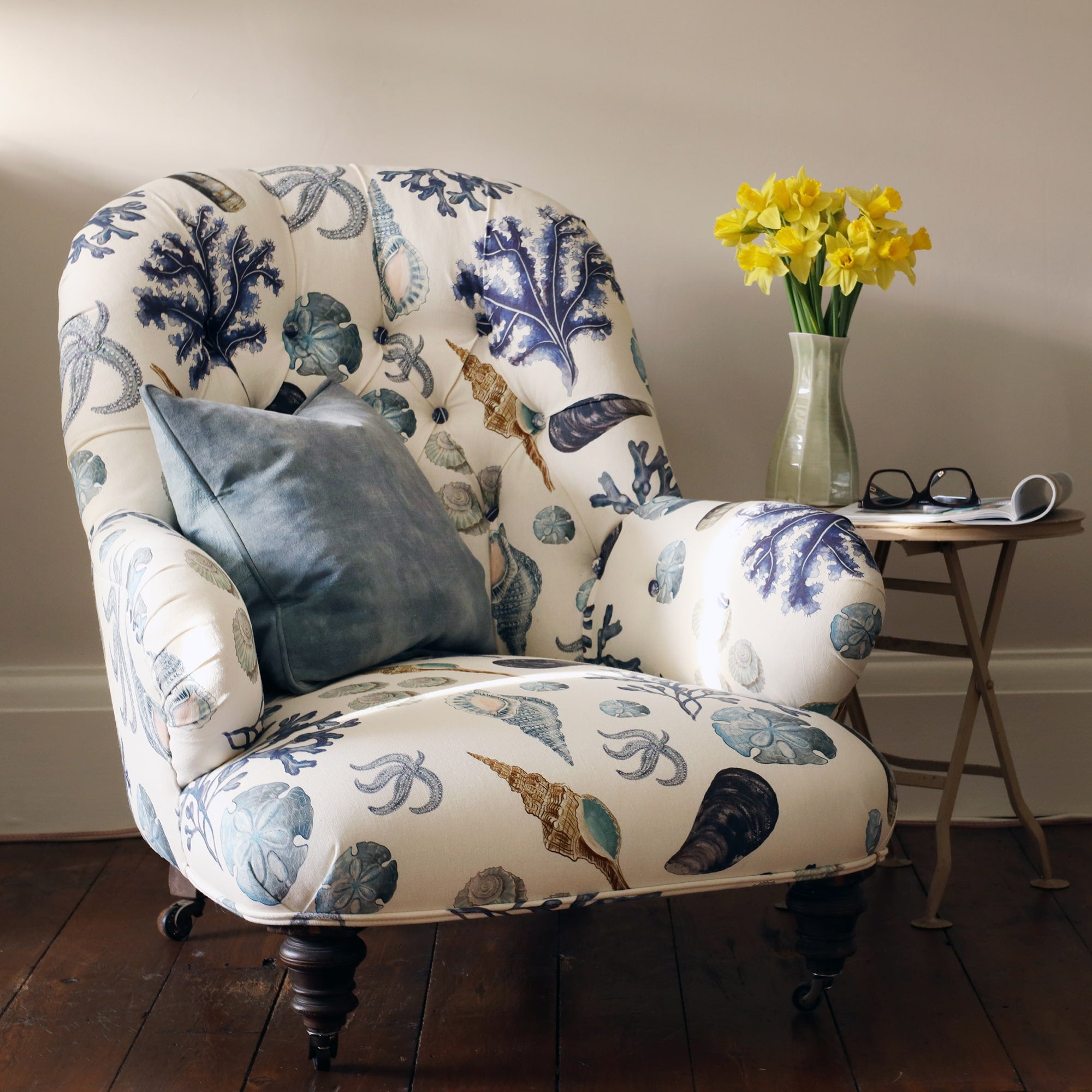 Front view of a re upholstered victorian button back armchair covered in a shell and seaweed fabric in blue tones on an off white background. The chair is sitting next to a small fold up table that has a vase of bright yellow daffodils, a folded magazine and a pair of reading glasses on it.