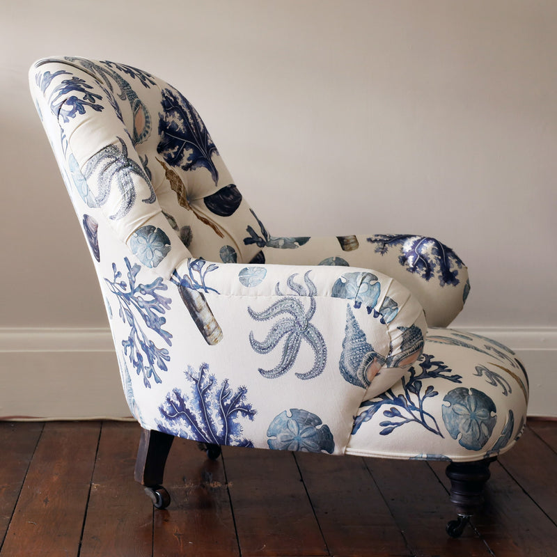 Side view of a re upholstered victorian button back armchair covered in a shell and seaweed fabric in blue tones on an off white background.