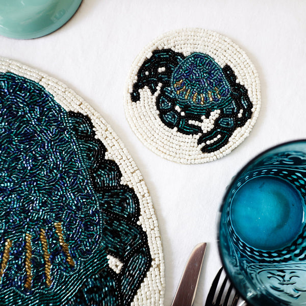 Handmade glass beaded coaster with a crab design  next to a matching placemat on a table setting