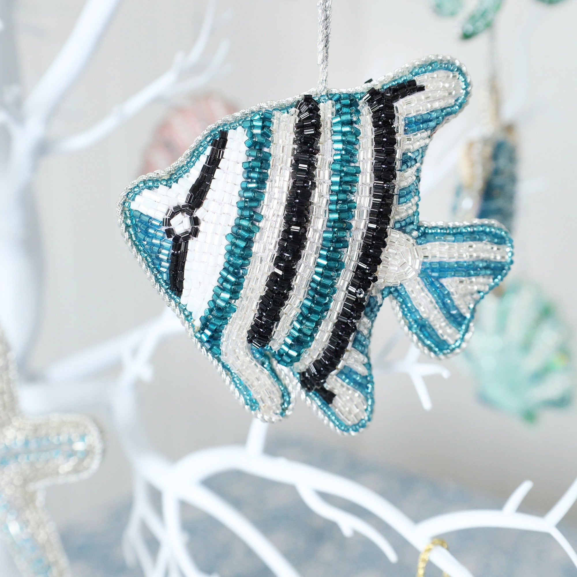 
The image shows a beaded fish ornament, hanging from a white tree branch. The fish features a striped pattern with beads in teal, black, white, and silver colors, giving it a shiny and intricate appearance. The tree in the background has other hanging decorations, and the setting suggests that it might be part of a festive or decorative display. The fish resembles an angelfish and the beads are arranged to create a textured, handcrafted look.