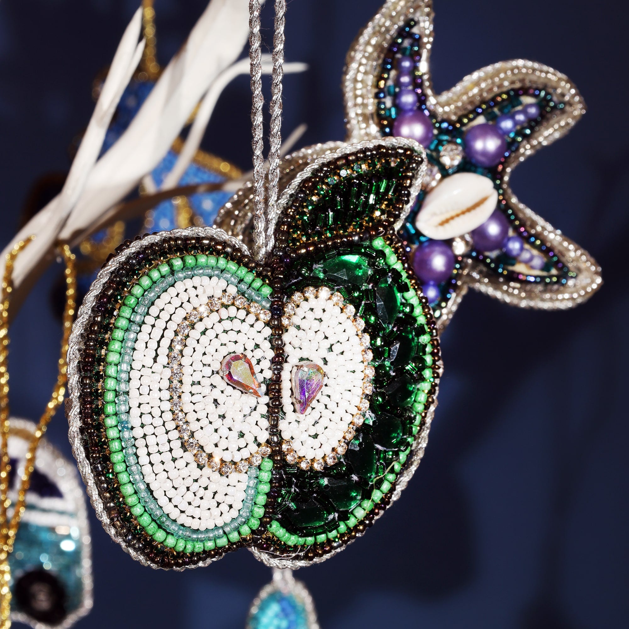 A close-up of a beaded apple-shaped Christmas ornament, intricately decorated with green, white, and black beads, along with sparkling accents. In the background, a starfish ornament made of purple, blue, and silver beads can be seen. Both ornaments are hanging from silver cords, set against a dark blue background, creating a festive and decorative scene.