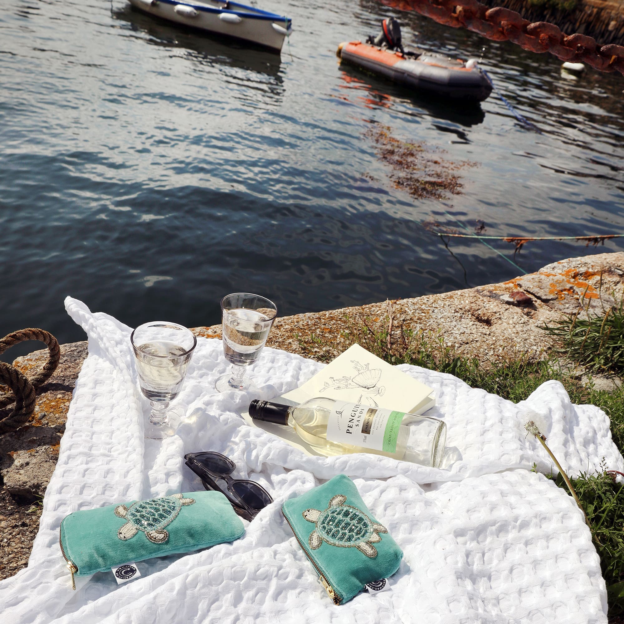 Picnic scene with white waffle blanket laid out on the quay with the water next to it. On the blanket there is a bottle of white wine, 2 glasses a notebook, sunglasses and an aqua velvet glasses case and coin purse both with a beaded turtle design on them.