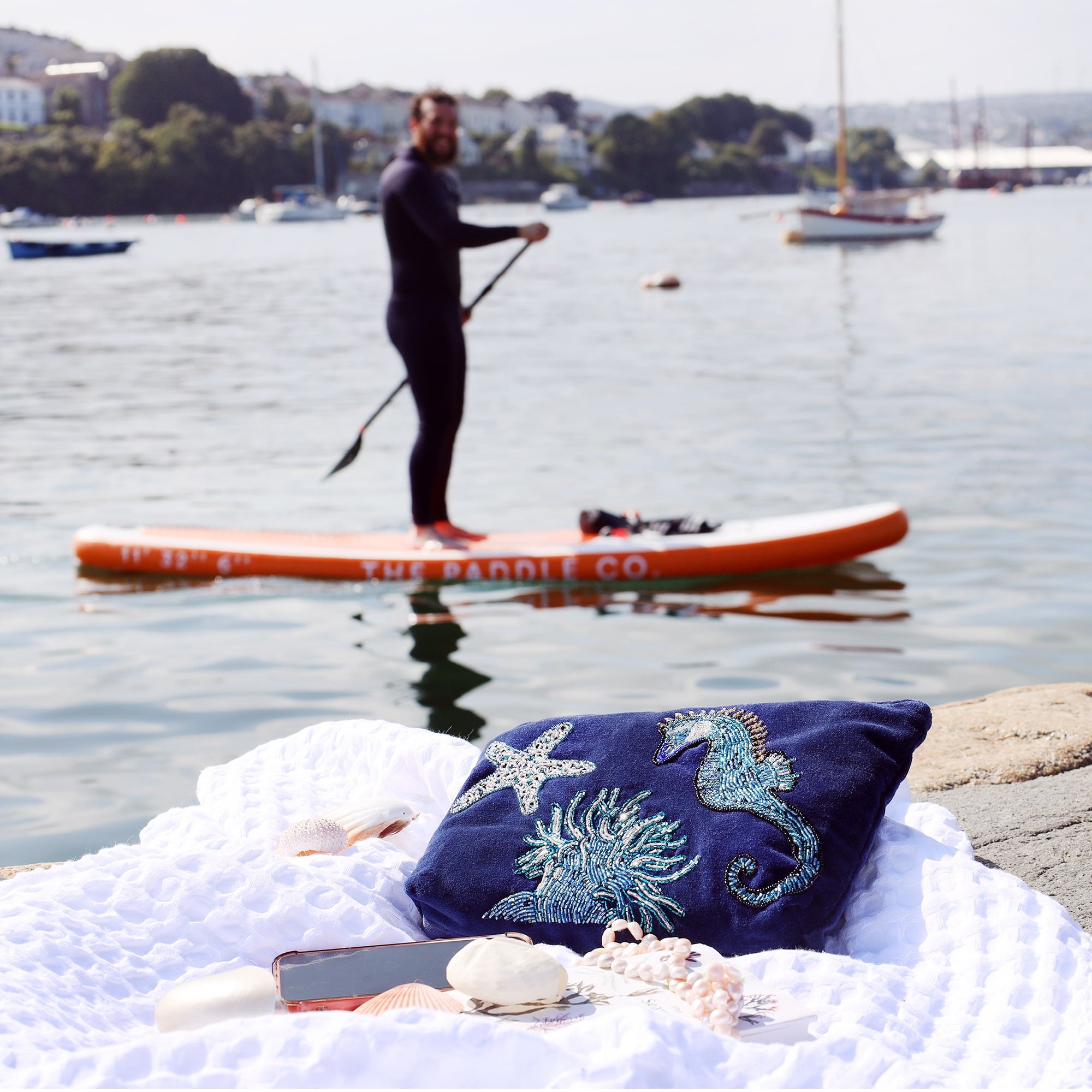 A navy velvet pouche decorated with a beaded seahorse, anemone and starfish laid on a white blanket with shells, mobile phone and pearls in front with a river scene of boats and a padle boarder going past in the background.