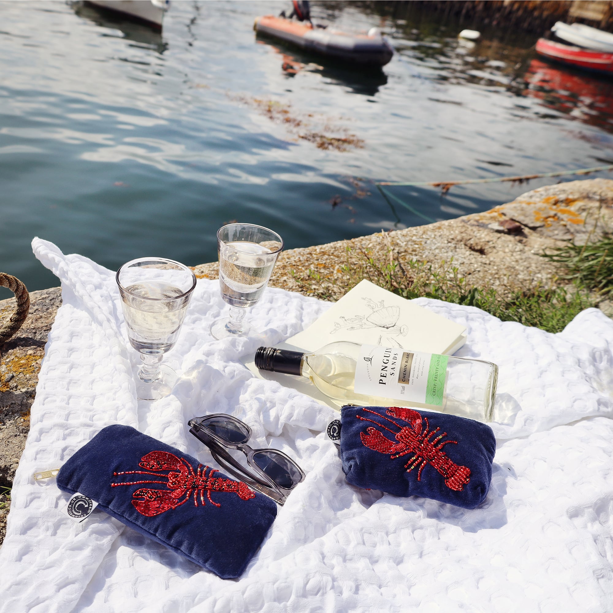 Picnic scene on the quay with water and boats in the background. On the white blanket is some wine and 2 wine glasses and a navy velvet glasses case and coin purse both with a red beaded lobster on the front.