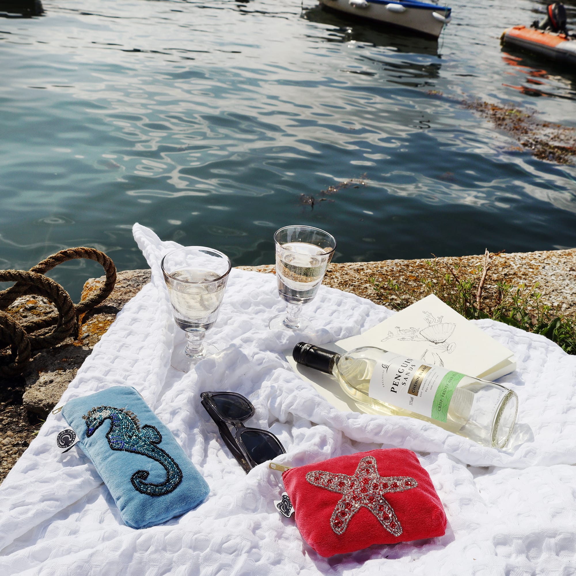 Picnic scene with white waffle blanket laid out on the quay with the water next to it. On the blanket there is a bottle of white wine, 2 glasses a notebook, sunglasses and an aqua velvet glasses case and coin purse and the other with a beaded starfish.