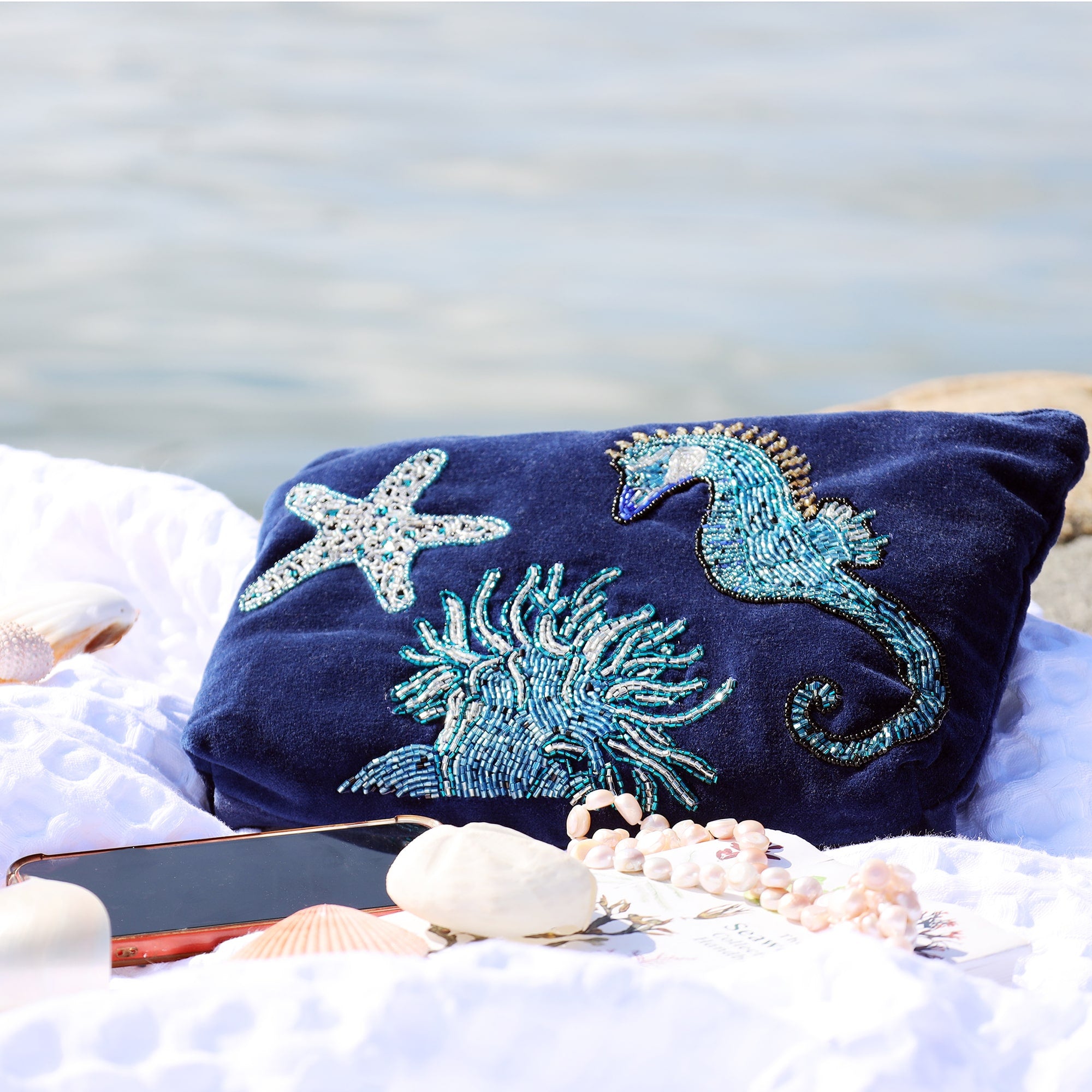 A navy velvet pouche decorated with a beaded seahorse, anemone and starfish laid on a white blanket with shells, mobile phone and pearls in front with the water in the background.