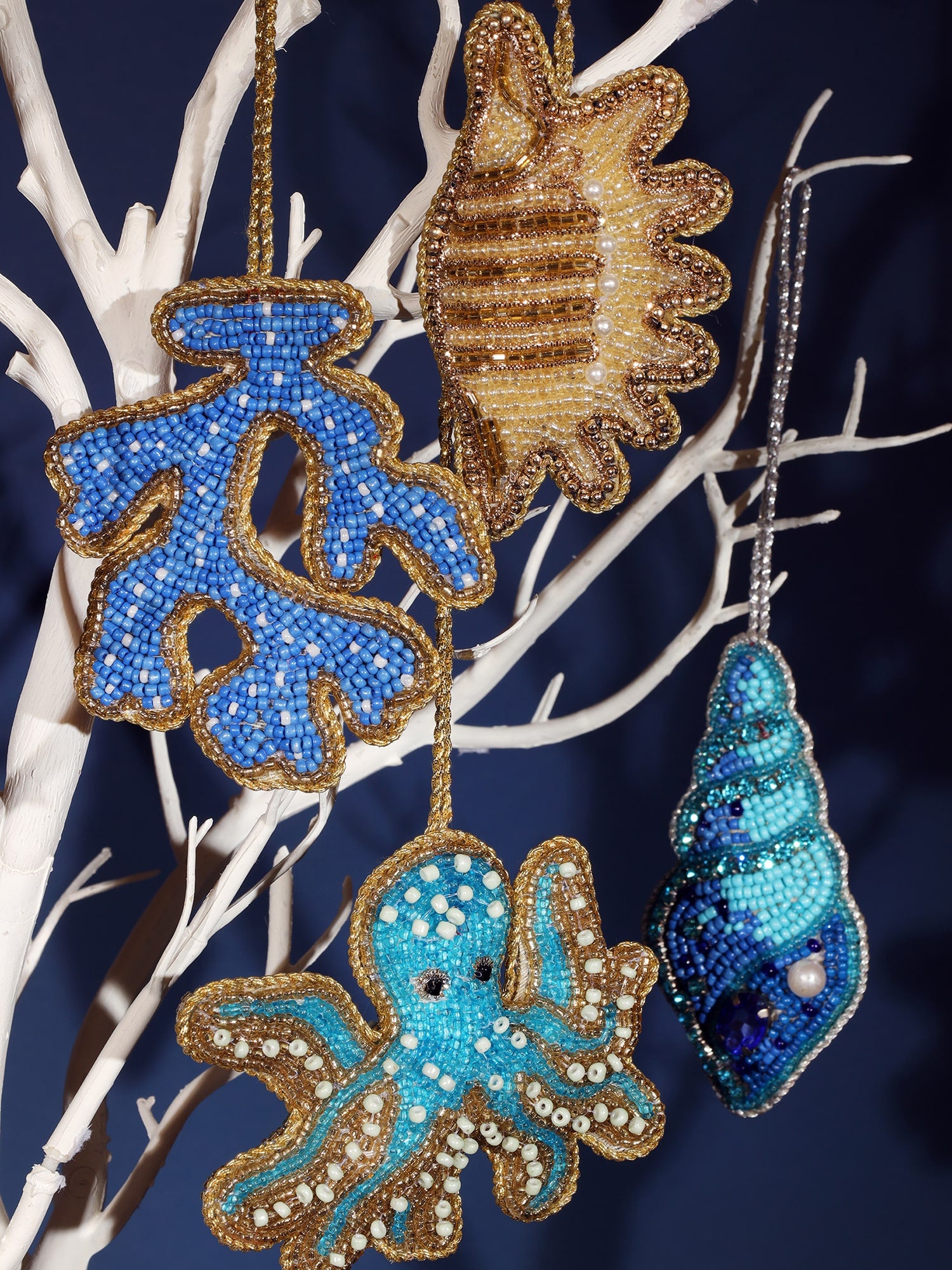 Close-up of colorful beaded Christmas ornaments hanging on a white decorative tree. The ornaments include a blue coral, a gold seashell, a blue octopus, and a blue seashell. Each ornament is intricately adorned with beads, pearls, and gold accents, creating a festive, ocean-inspired theme. The display is set against a dark blue background, highlighting the vibrant colors of the decorations.