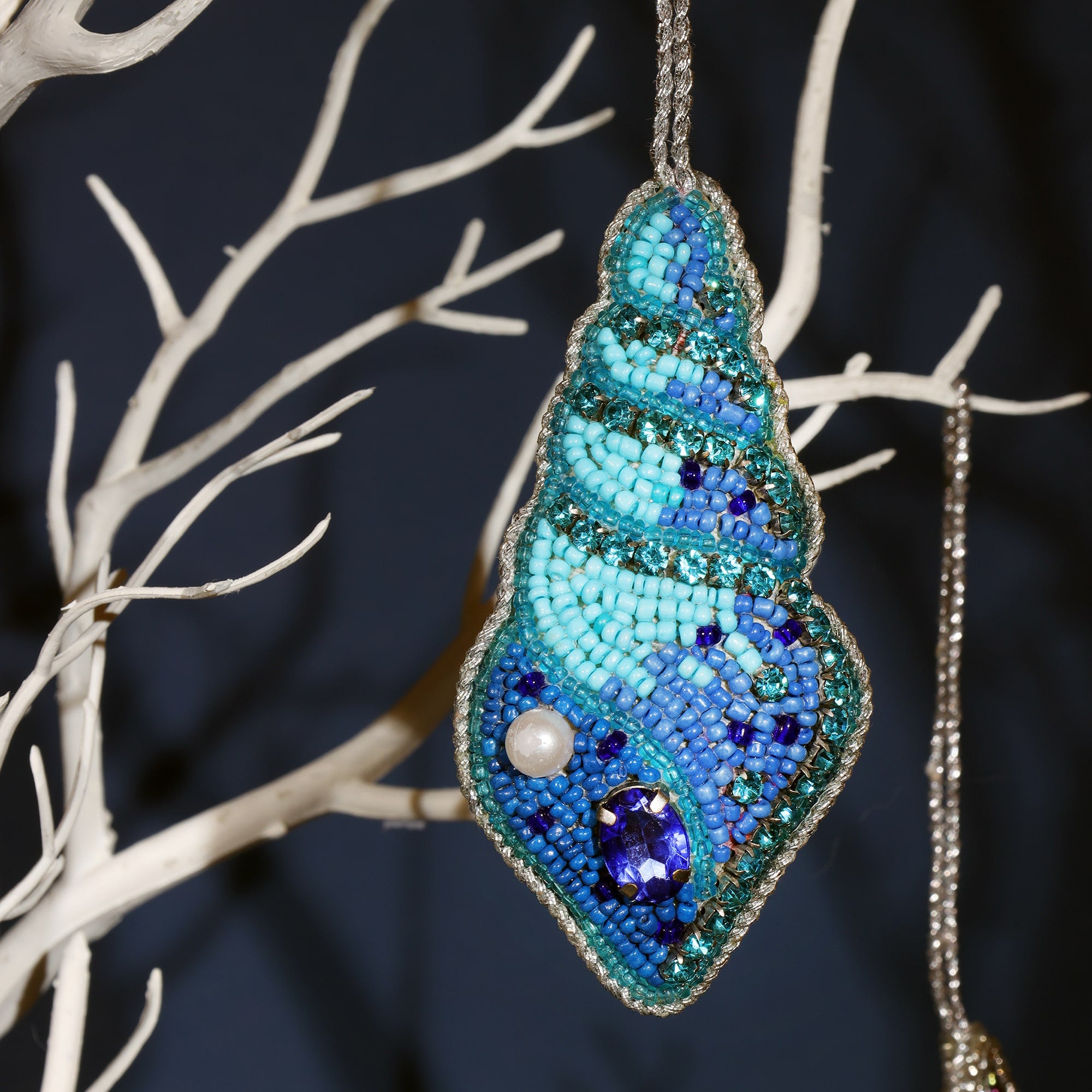 A close-up of a beautiful beaded seashell ornament hanging on a white decorative tree. The ornament is intricately crafted with shades of blue and turquoise beads, accented with sparkling rhinestones and a small pearl. A silver border outlines the shell, giving it a shimmering, elegant look.