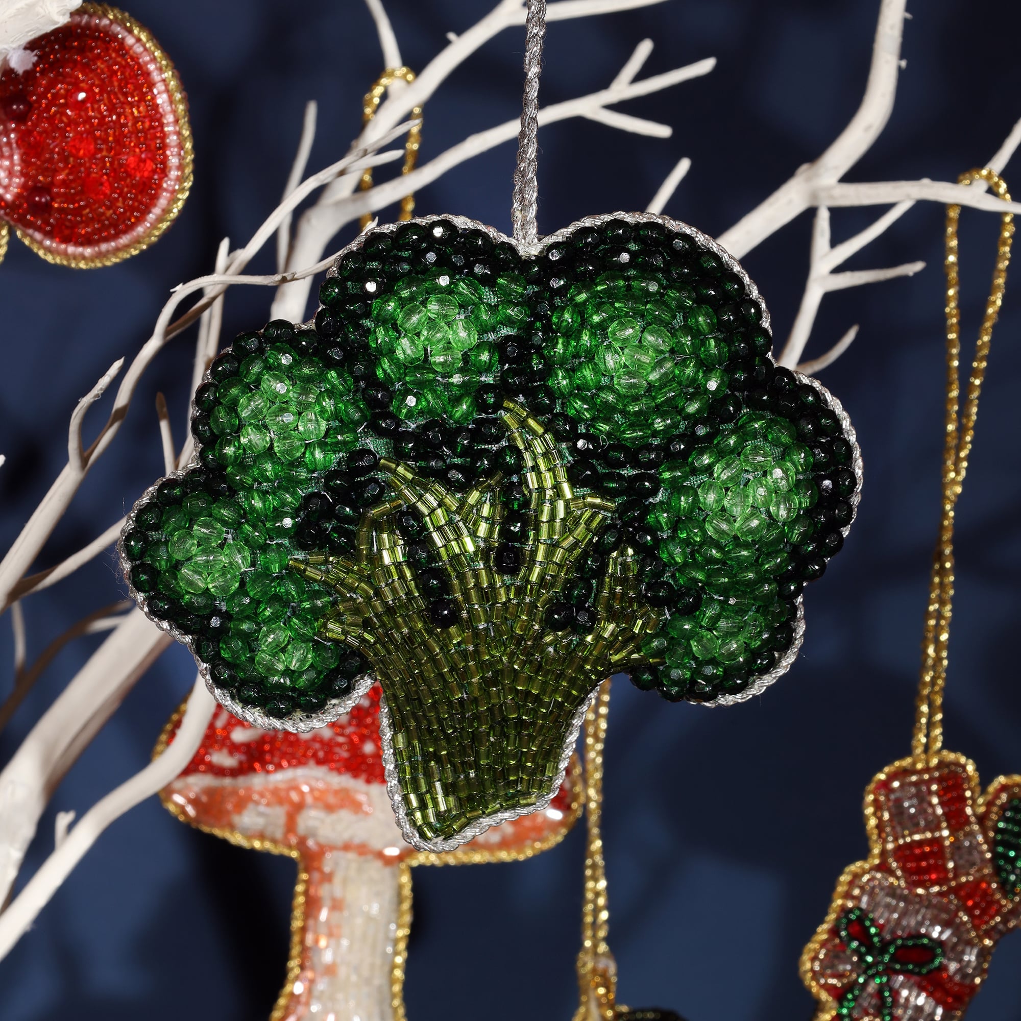 A close-up of a beaded broccoli ornament hanging on a white decorative tree. The ornament features vibrant green beads for the florets and lighter green and black beads for the stem, with a silver beaded outline. The detailed craftsmanship gives the broccoli a realistic yet whimsical appearance.