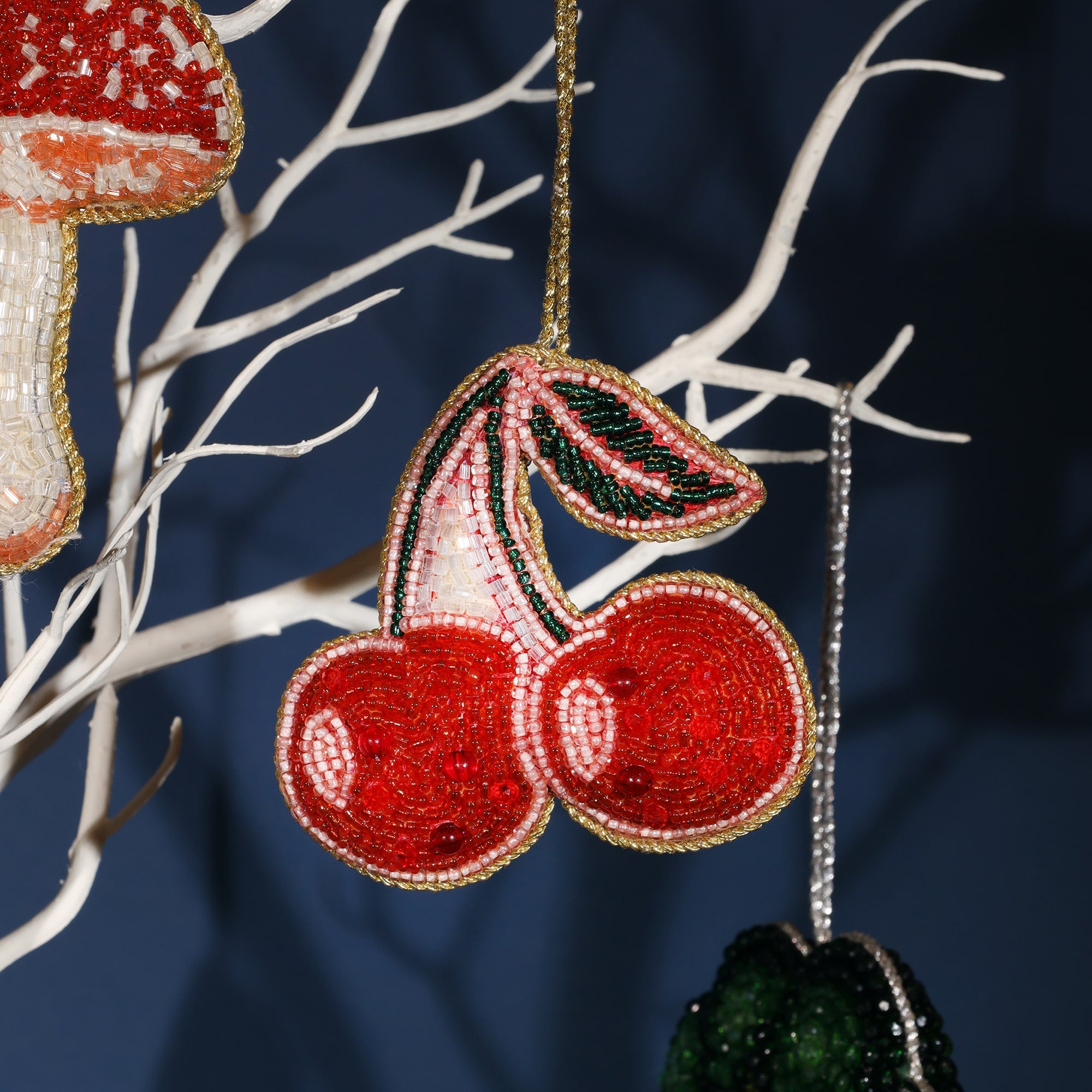 Close-up of a vibrant beaded ornament in the shape of two red cherries with green leaves, hanging from a white decorative tree branch. The cherries are intricately crafted with red, pink, and green beads, and outlined in gold beading, giving them a festive and eye-catching appearance. In the background, part of a mushroom-shaped beaded ornament is visible.