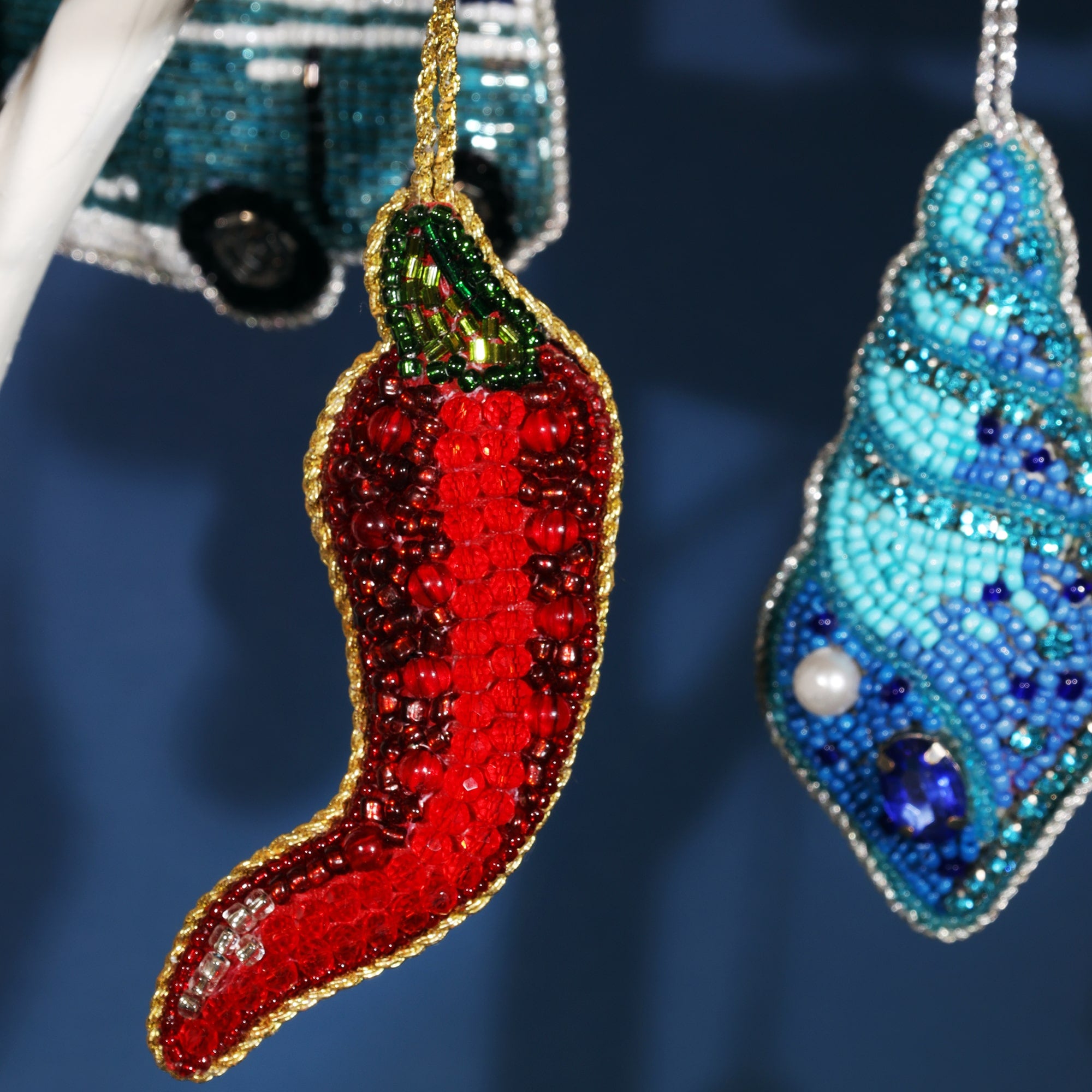 Close-up of a beaded chili pepper ornament. The pepper is bright red with detailed beadwork, accented by a green stem and outlined in gold beads. It hangs against a dark blue background, adding a vibrant pop of color to the scene.