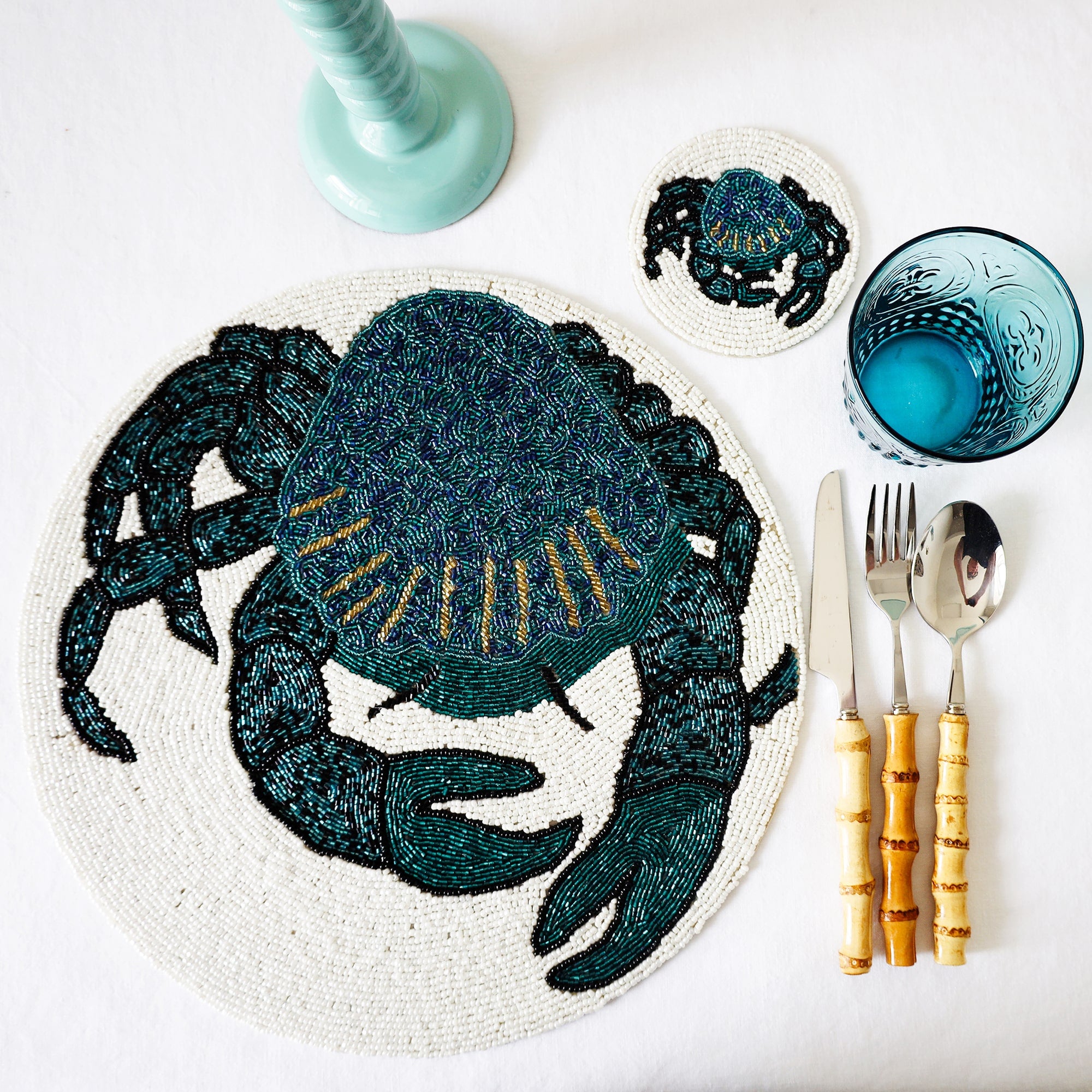Handmade glass beaded coaster with a Crab design next to a matching placemat on a table setting with glasses,cutlery and a candlestick
