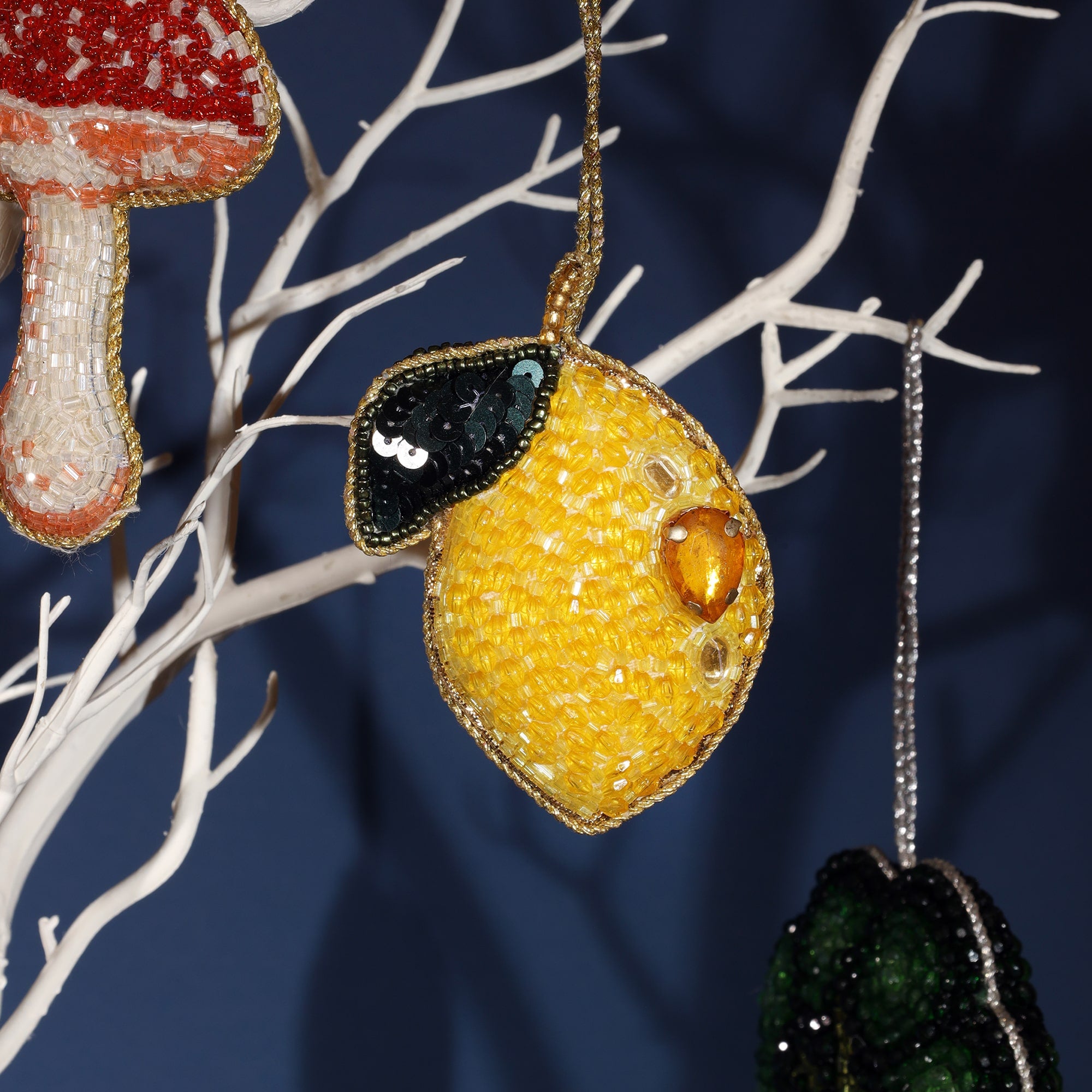 A close-up of a vibrant beaded lemon ornament hanging on a white decorative tree branch. The lemon is crafted with bright yellow beads and features a glossy green sequin leaf, along with a golden teardrop-shaped gem. The intricate beadwork adds a shiny texture, making the lemon stand out as a bold and playful decoration.